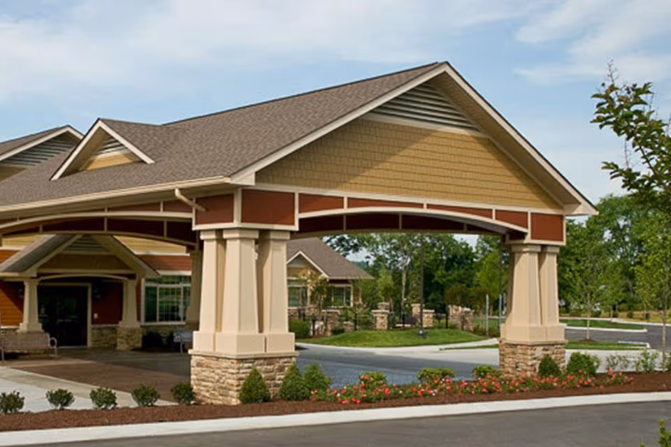 Front entrance of a senior living facility showing a covered porte-cochere with columns, stone accents, and a landscaped driveway.
