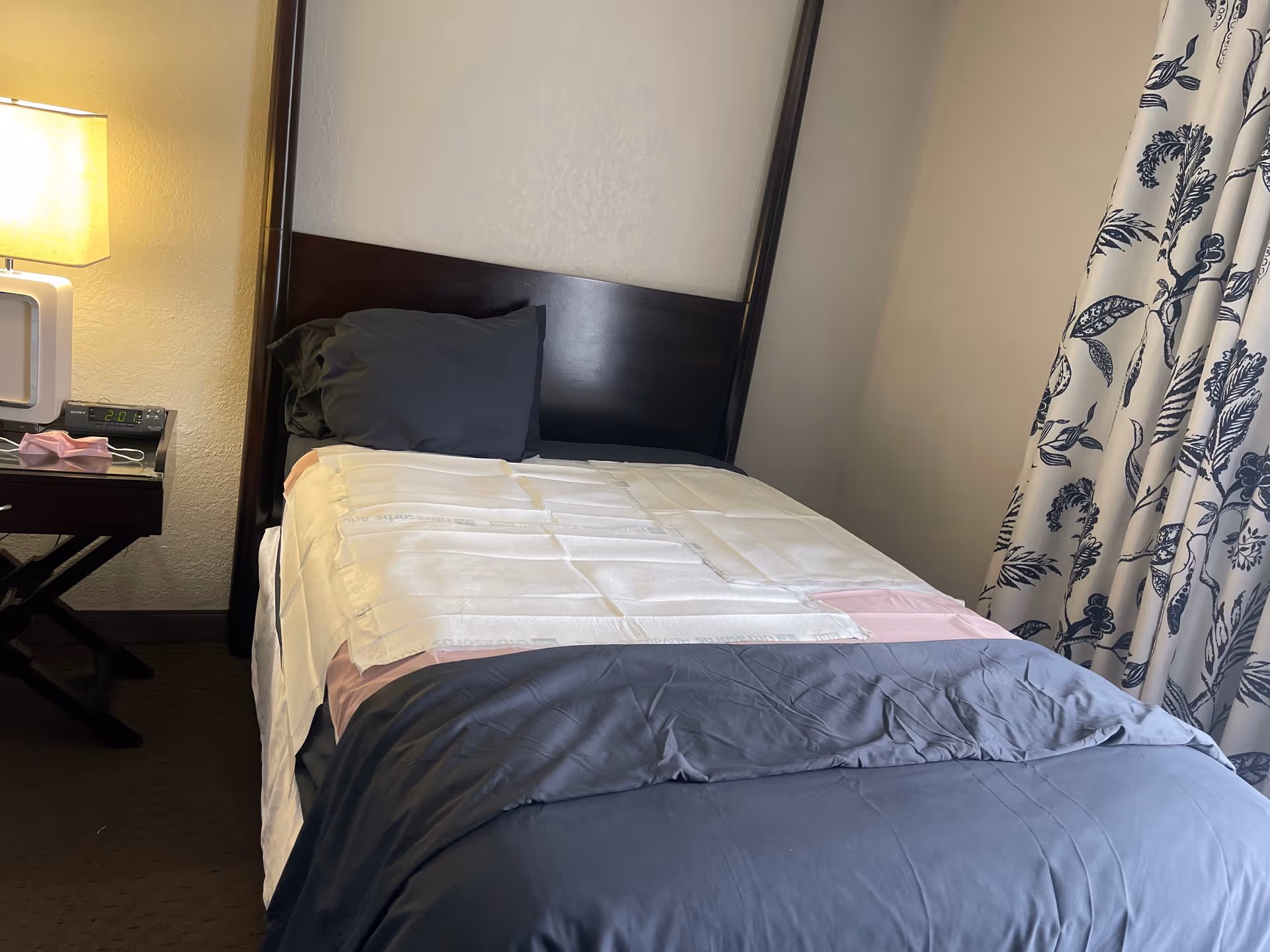 A single bed in a small bedroom with a lamp, nightstand, and patterned curtains.