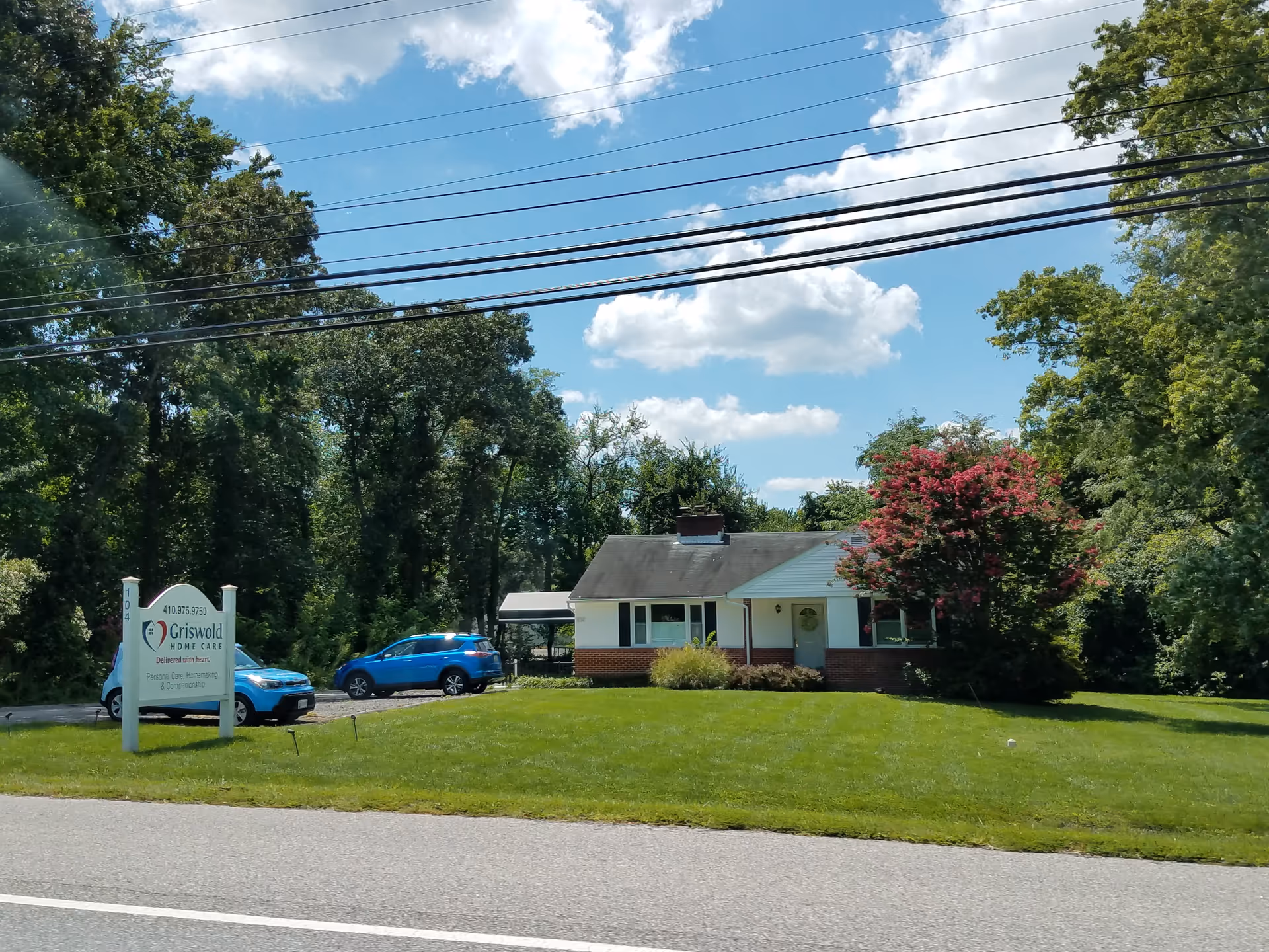 Single-story building with white trim, green lawn, sign, and a blue car parked in driveway.