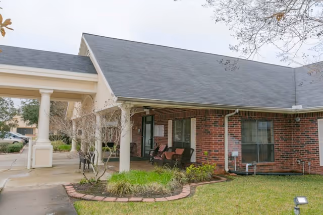 Exterior view of a brick building with a covered entrance supported by white columns. There are two chairs with cushions on the porch, some small plants and grass in the foreground, and a tree with bare branches. The sky is overcast.