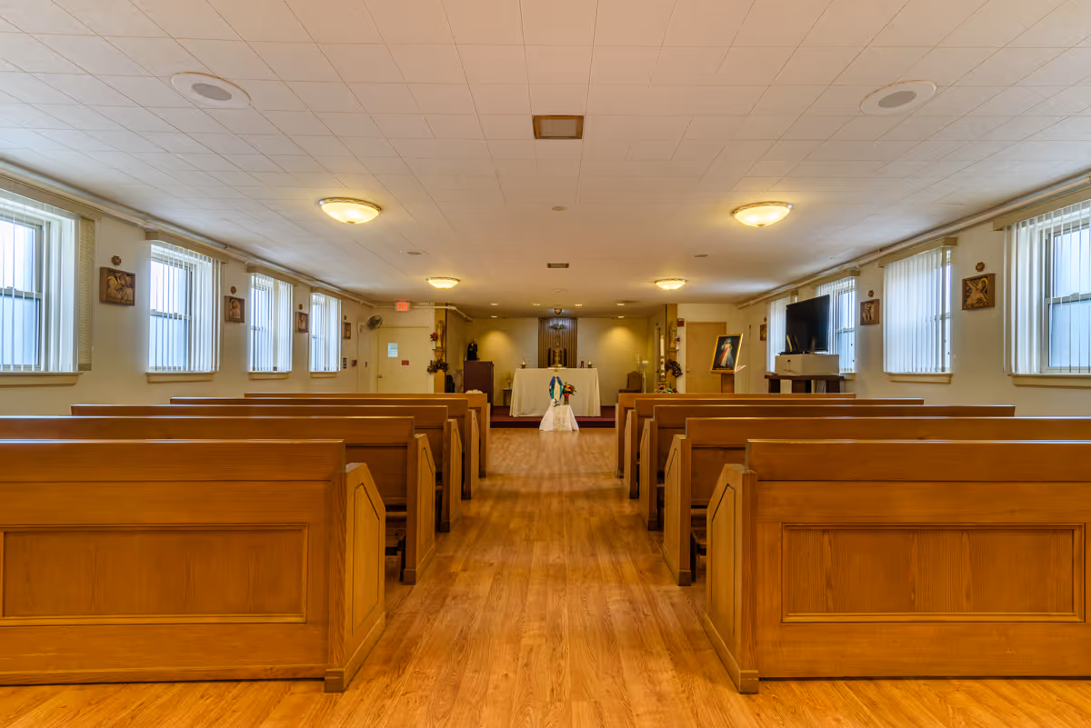 Interior view of a chapel with wooden pews arranged in rows on both sides of a central aisle leading to an altar at the front. The room has multiple windows with vertical blinds, religious artwork on the walls, and ceiling lights providing illumination.