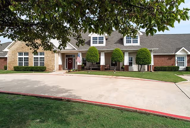 Front exterior view of a single-story residential building with a stone and brick facade, a covered porch with seating, neatly trimmed bushes, an American flag near the entrance, and a curved driveway bordered by grass and a tree overhead.