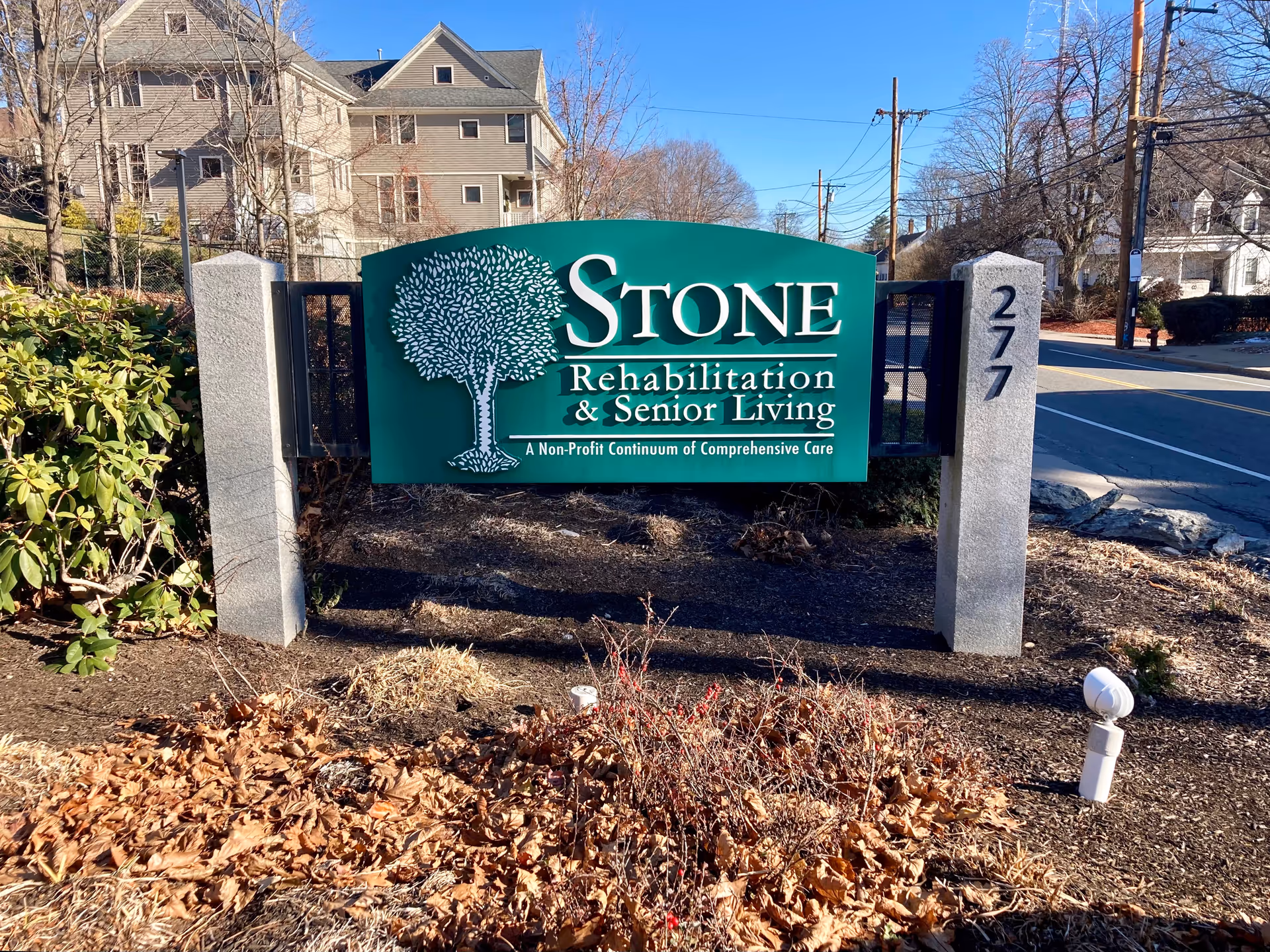 Green entrance sign reading "Stone Rehabilitation & Senior Living" mounted between stone posts with buildings and a road in the background.