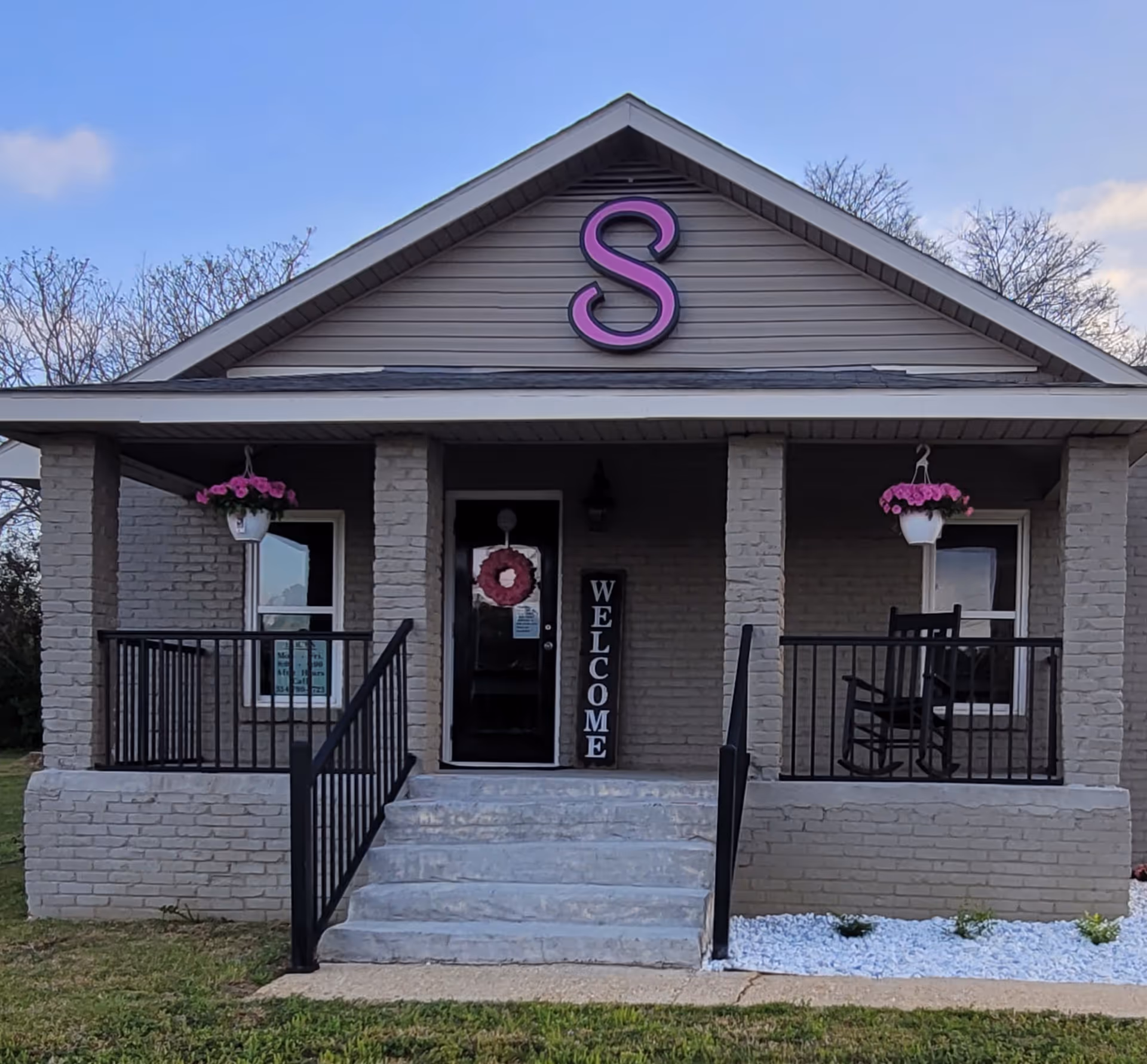 Front view of a brick senior facility with a porch, welcome sign, and hanging planters.