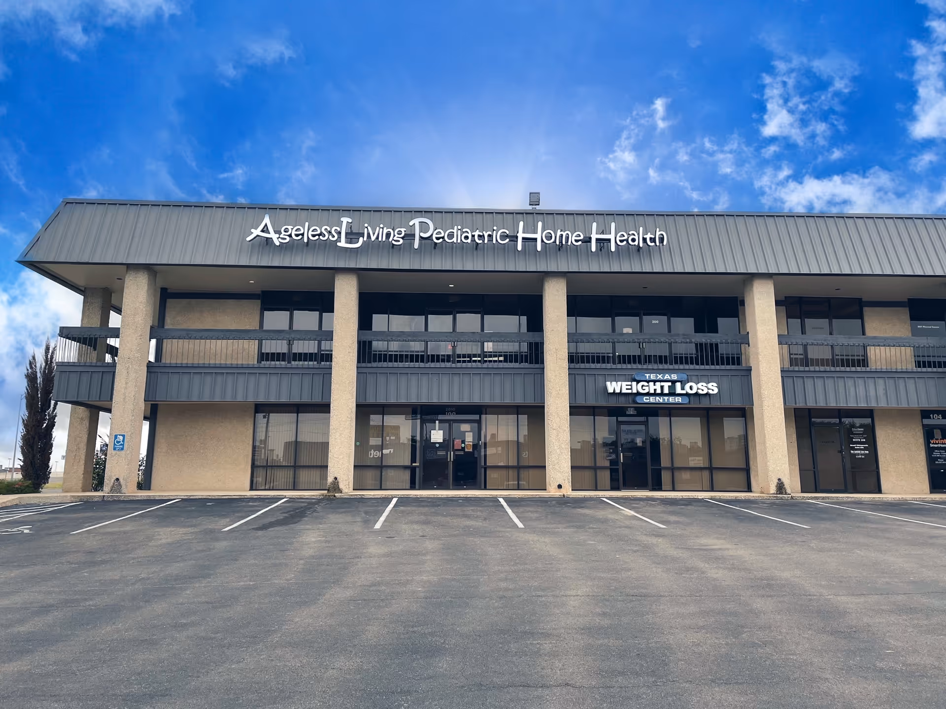 Two-story commercial building facade featuring Ageless Living Pediatric Home Health sign and empty parking.