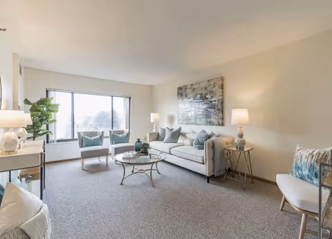 A bright and spacious living room with a large window letting in natural light. The room features a white sofa with blue and gray cushions, two matching armchairs, a glass coffee table with decorative items, a side table with a lamp, and a large abstract painting on the wall. The carpet is light gray, and there is a potted plant near the window.