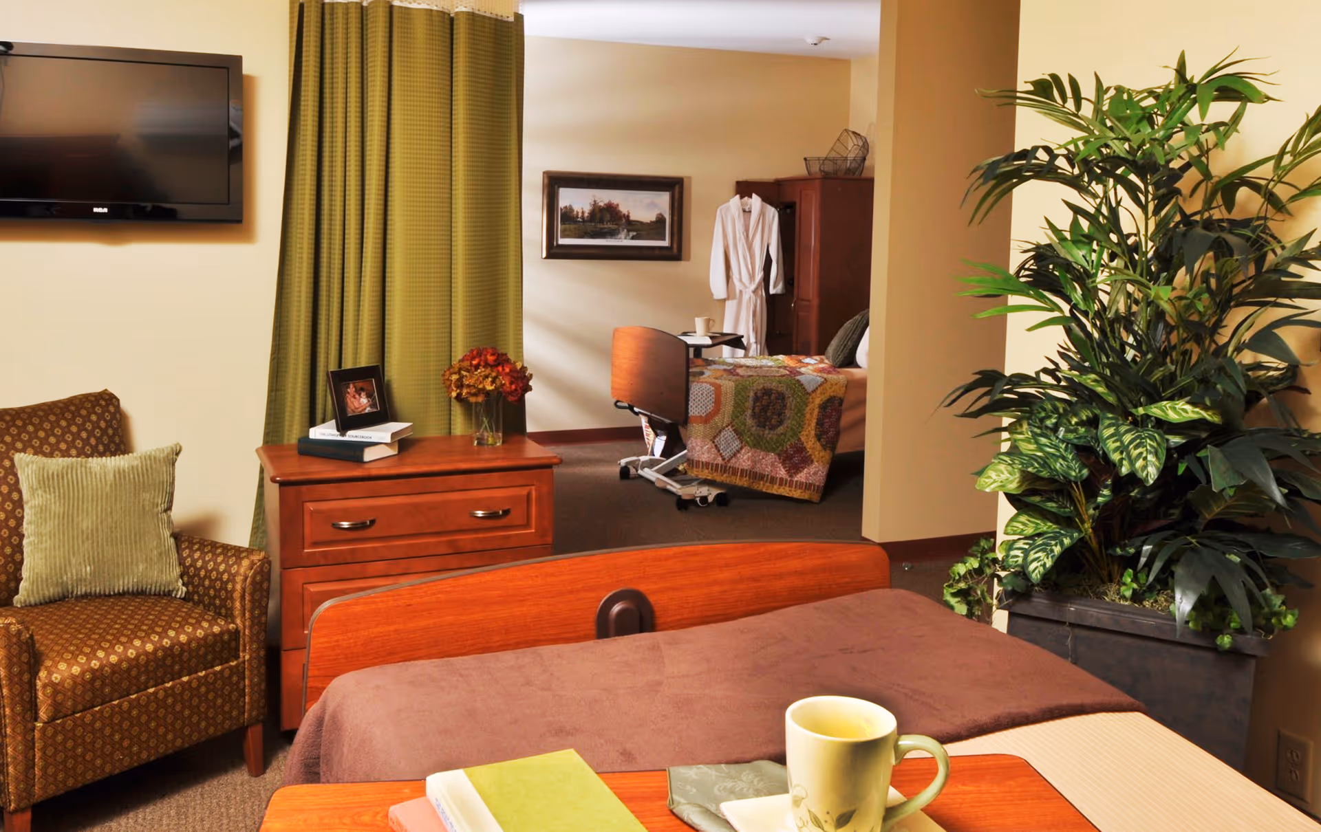 A cozy senior living bedroom with a brown bedspread, a wooden headboard, and a bedside table holding a cup and books. To the left, there is a patterned armchair with a green pillow, a wooden dresser with a framed photo, books, and a vase of flowers. A flat-screen TV is mounted on the wall above green curtains. In the background, another bedroom area is visible with a bed covered by a colorful quilt, a white robe hanging on a wardrobe, and a framed picture on the wall. A large potted plant is on the right side of the image.