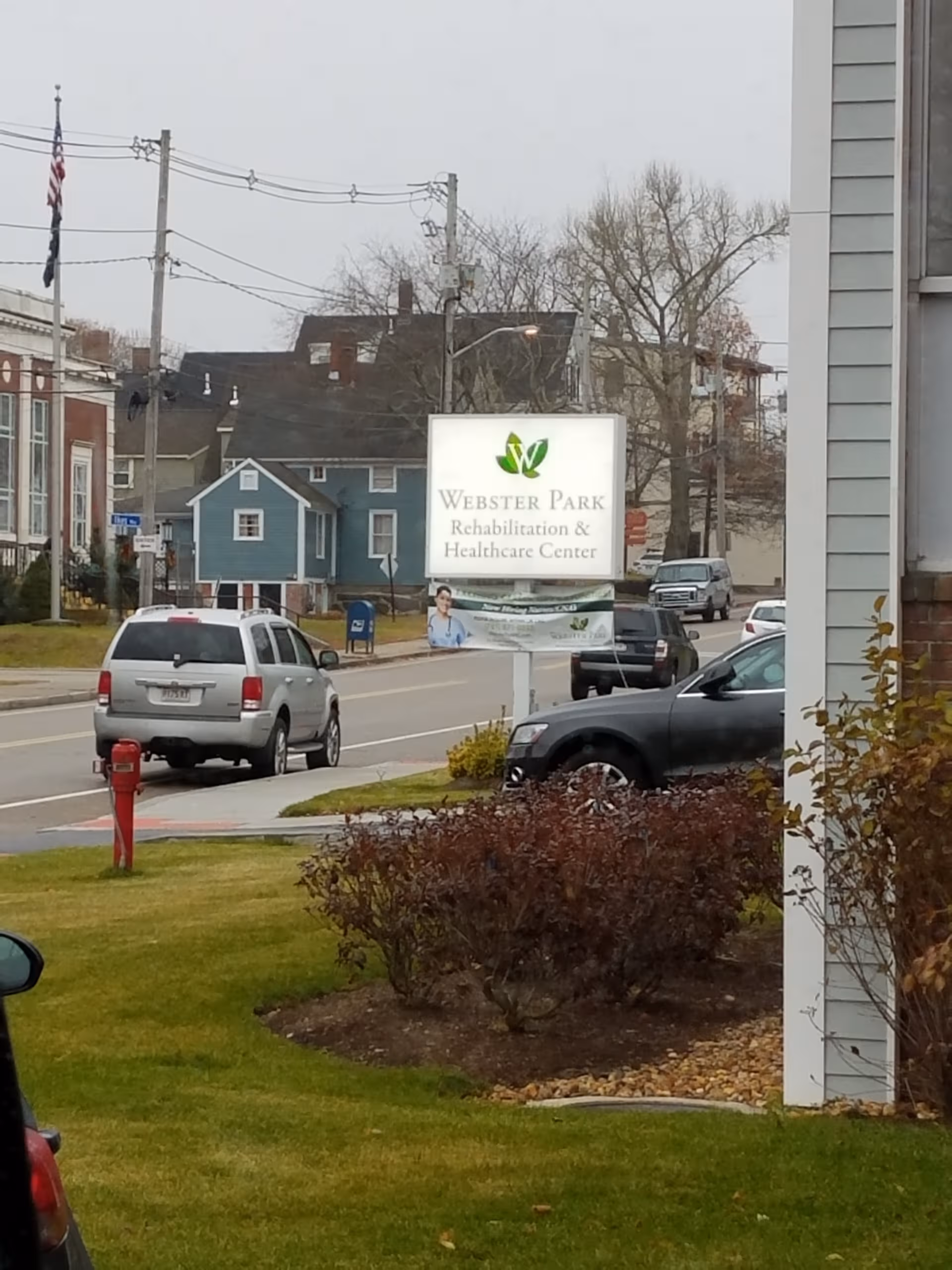 Street view of Webster Park Rehabilitation & Healthcare Center sign with parked cars and houses.