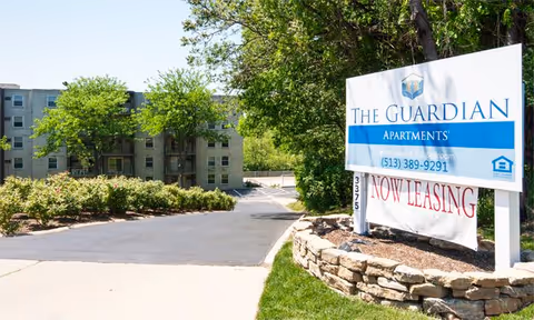 Entrance to The Guardian Apartments with a large sign displaying the facility name, phone number, and a 'Now Leasing' banner. The driveway leads to a multi-story apartment building surrounded by trees and greenery.