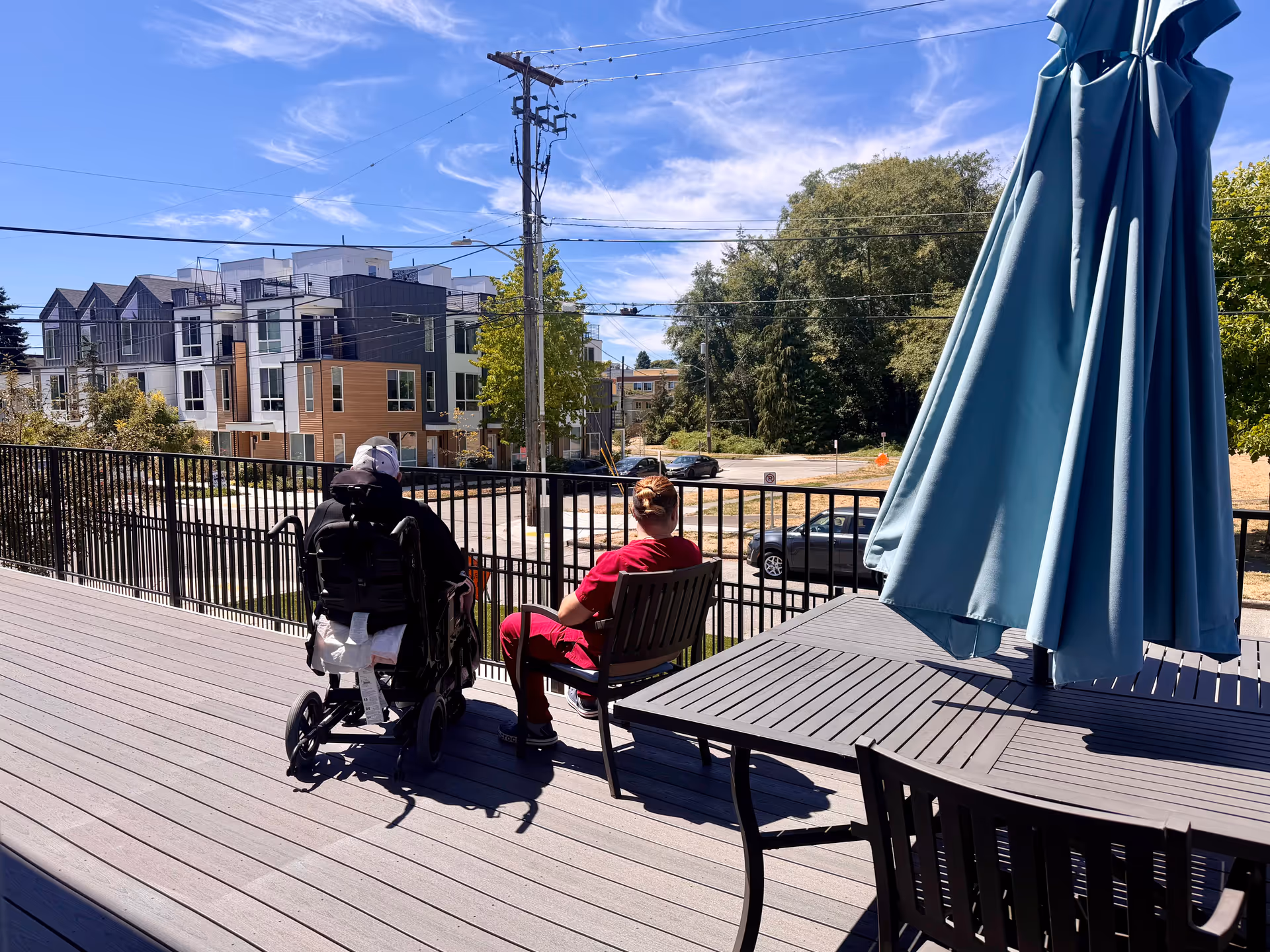 An outdoor patio deck with two people seated, wheelchair user, umbrella, and modern building in background.
