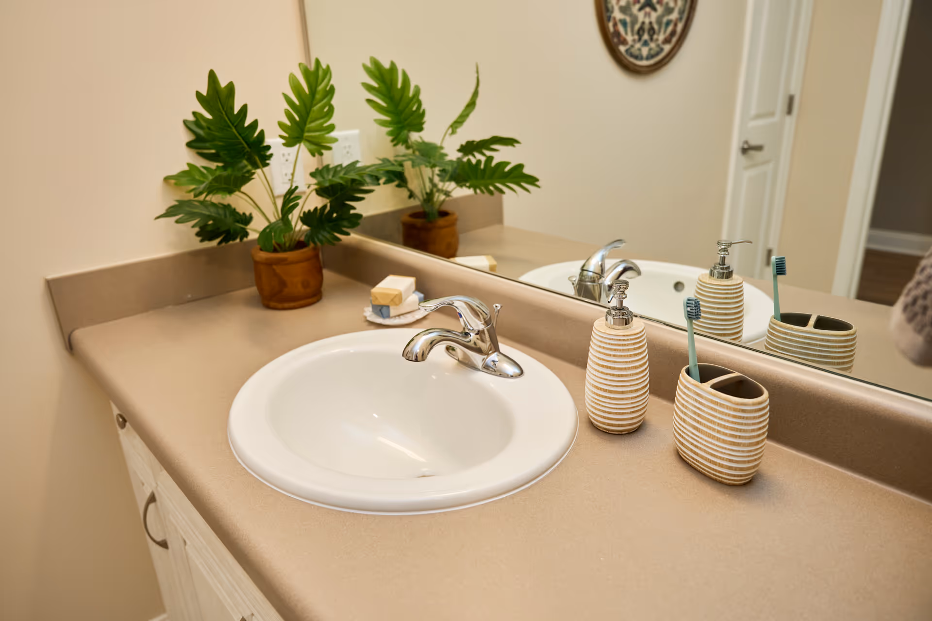 Bathroom sink with beige countertop, mirror, soap dispensers, and potted plants.