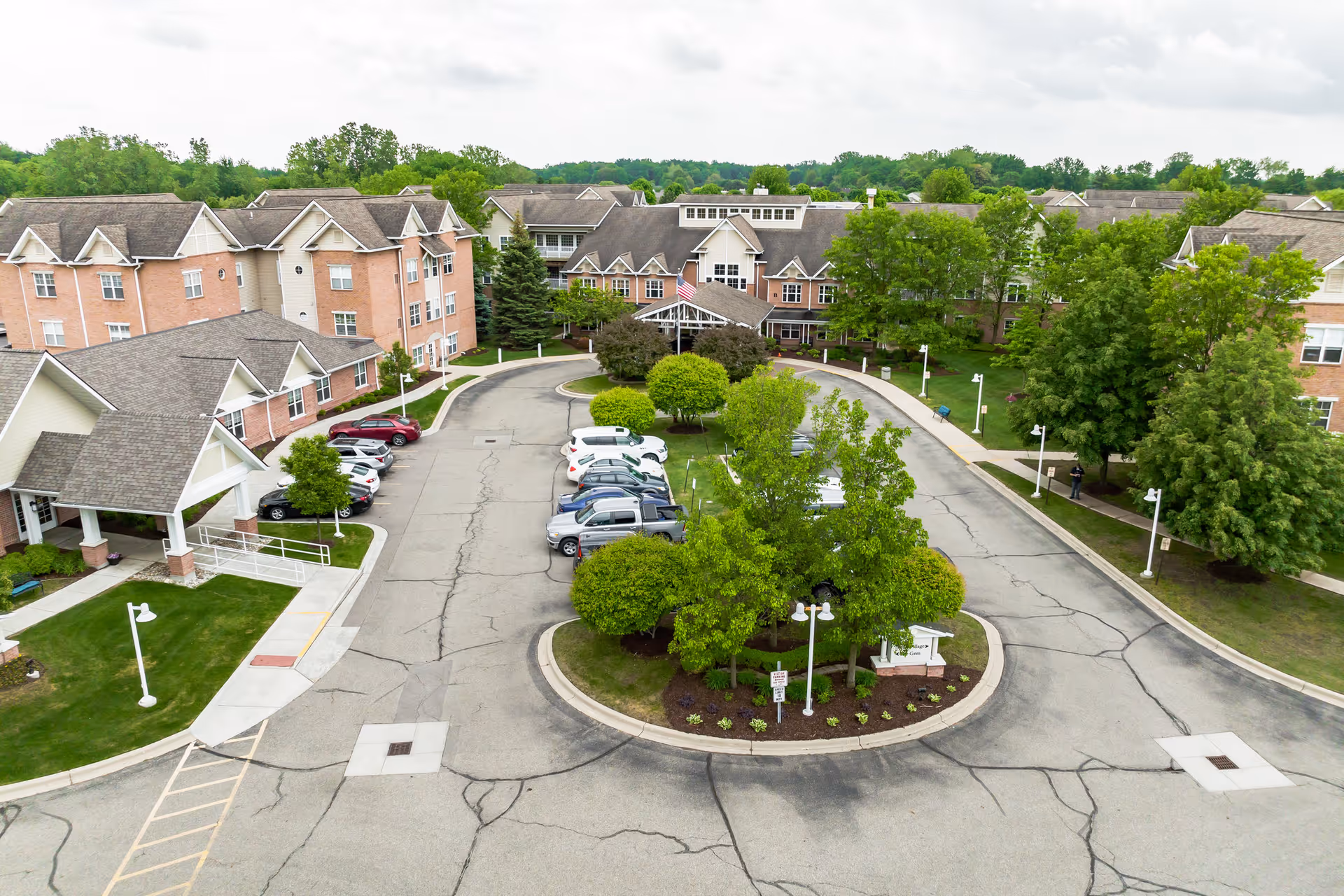 Aerial view of a senior living complex showing a circular driveway with parked cars and multi-story brick buildings set among trees and landscaping.