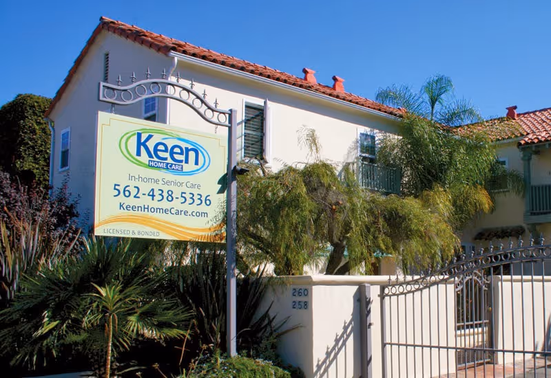 Exterior view of Keen Home Care building with sign, gate, and palm trees.