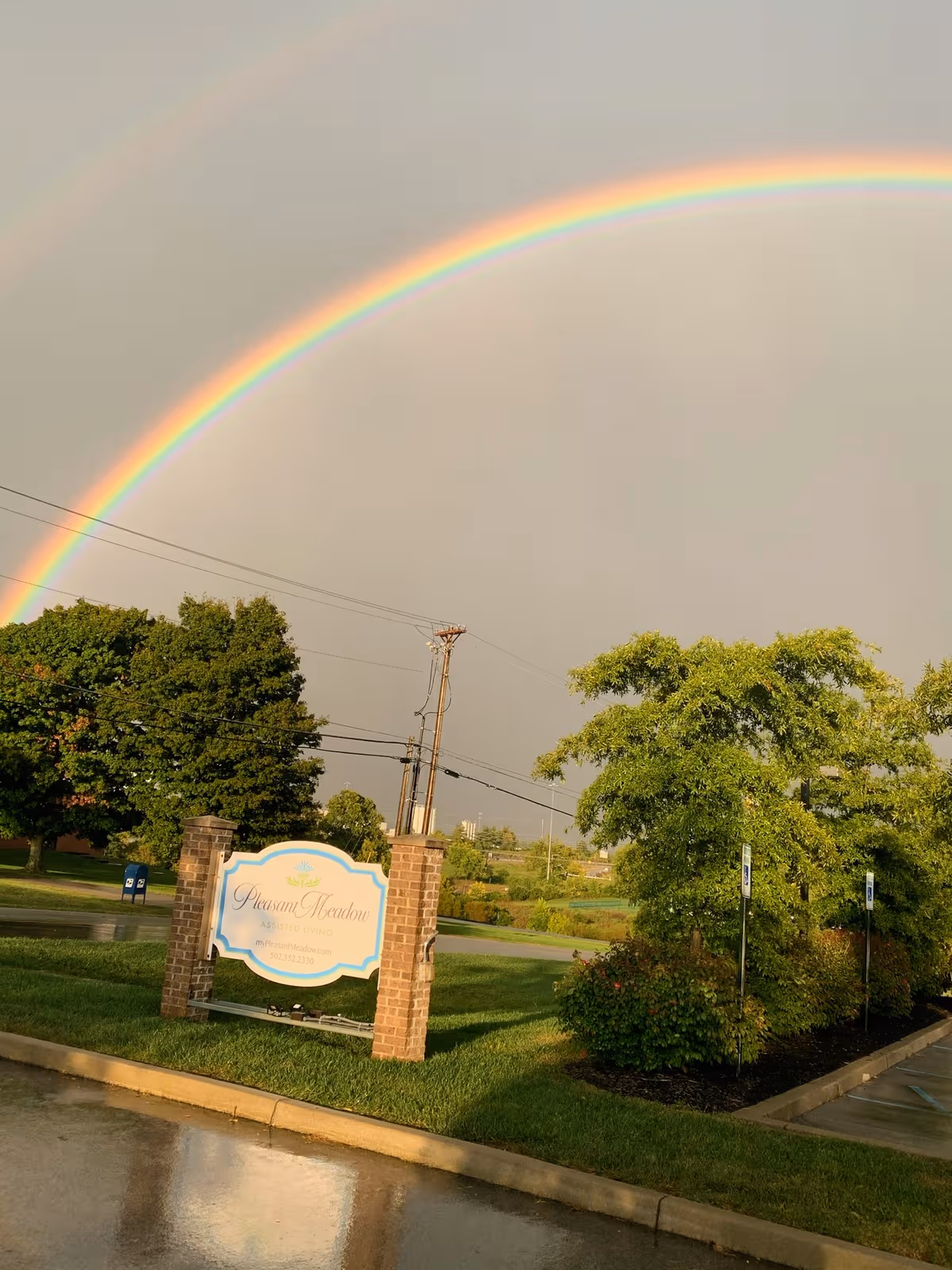 Double rainbow arches over Pleasant Meadow Assisted Living entrance with brick sign and trees.
