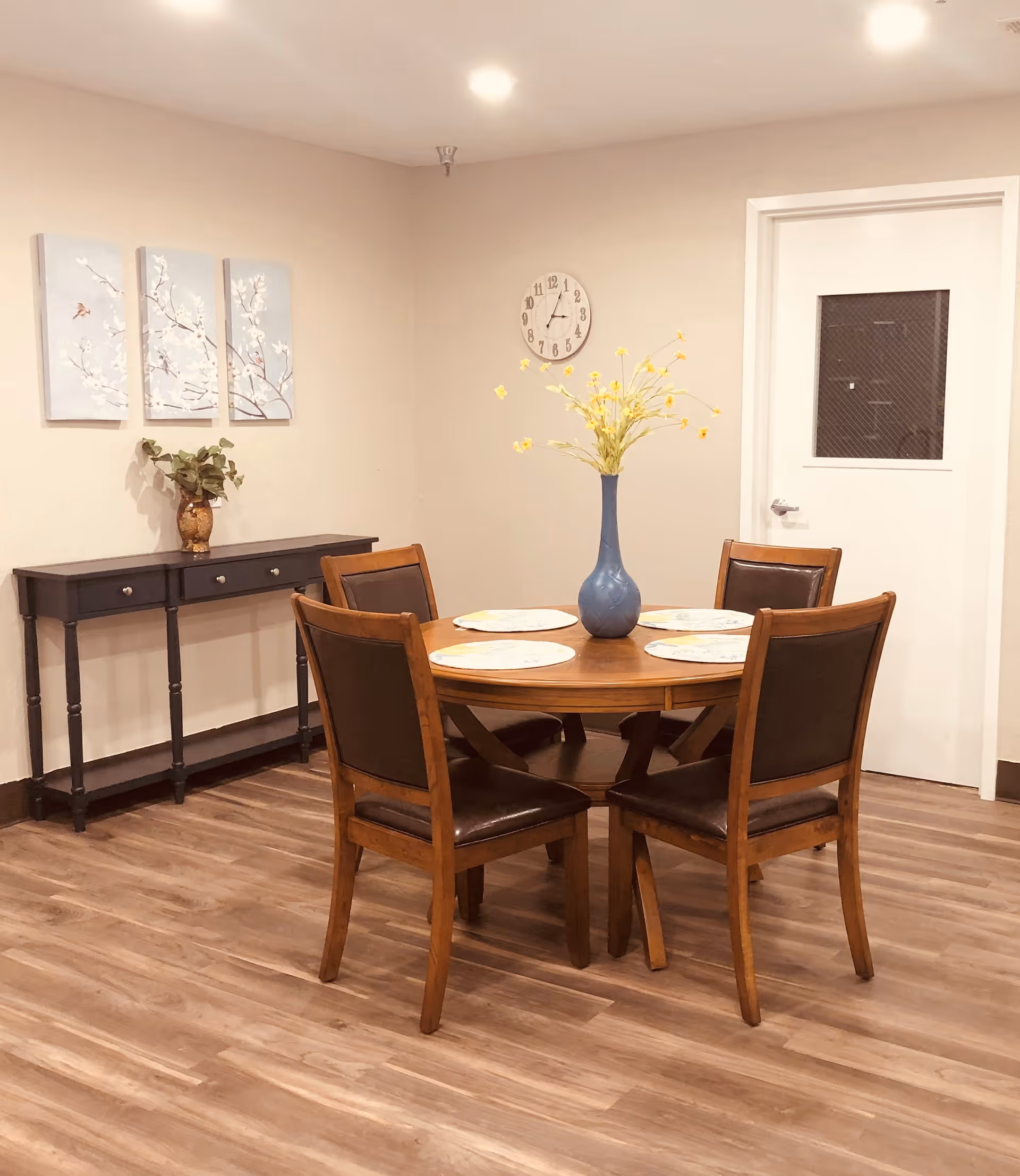 A dining area with a round wooden table set with four placemats and a blue vase with yellow flowers in the center. Four wooden chairs with dark cushioned seats surround the table. Against the wall is a narrow black console table with a small plant in a vase. Above the console table are three decorative wall panels featuring a floral design. A clock is mounted on the wall near a white door with a small window.