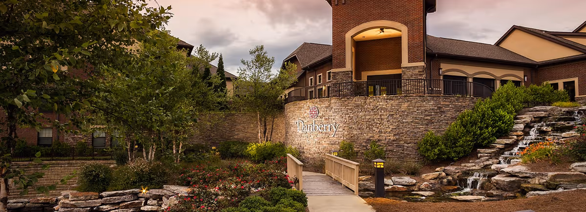 Front exterior of Danberry at Inverness showing a stone entrance wall with the Danberry sign, a small bridge over landscaped ponds and a waterfall.