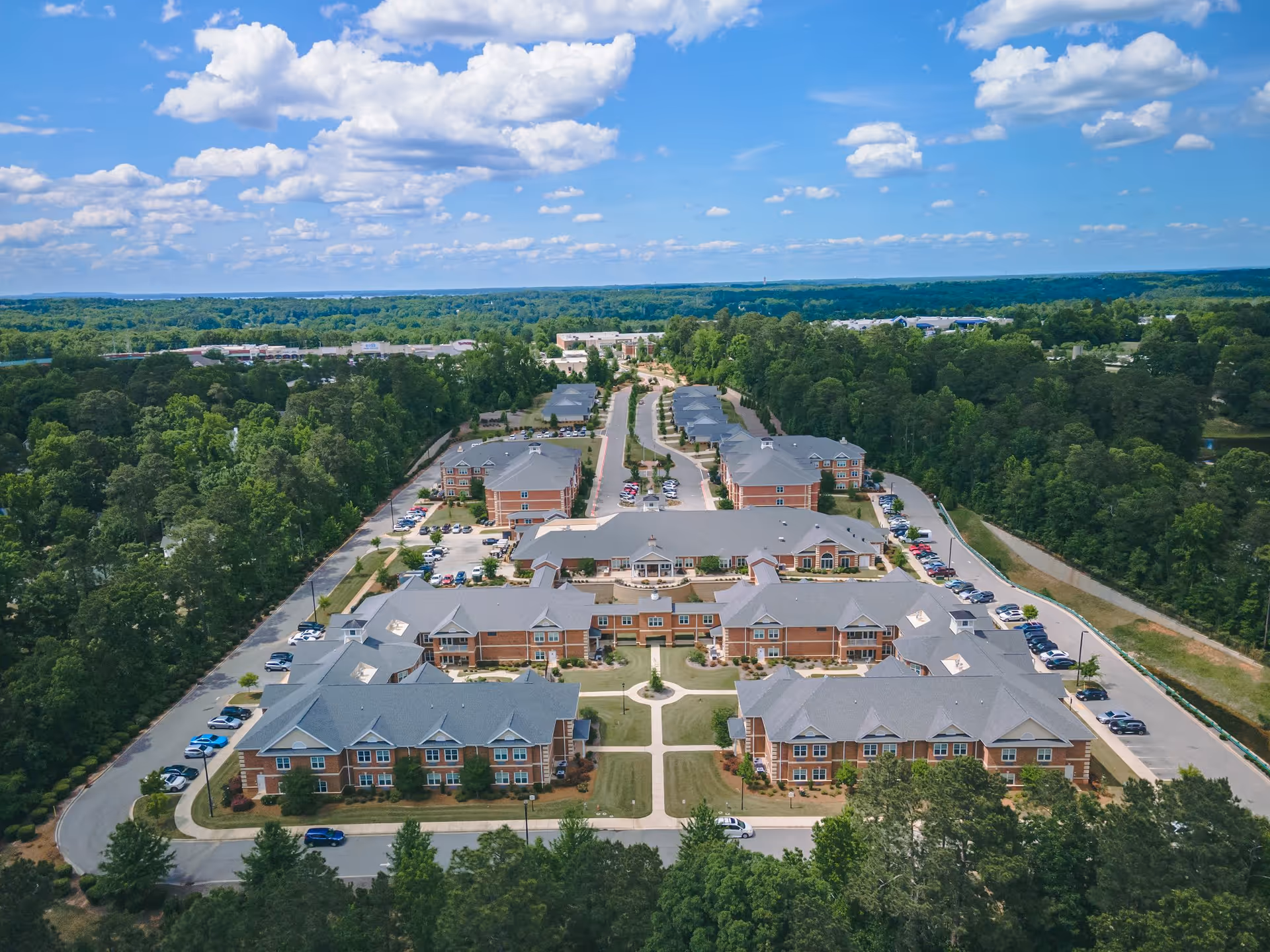 Aerial view of Wellmore of Lexington senior living facility showing multiple brick buildings with gray roofs arranged around landscaped green lawns and parking areas, surrounded by dense trees under a partly cloudy blue sky.