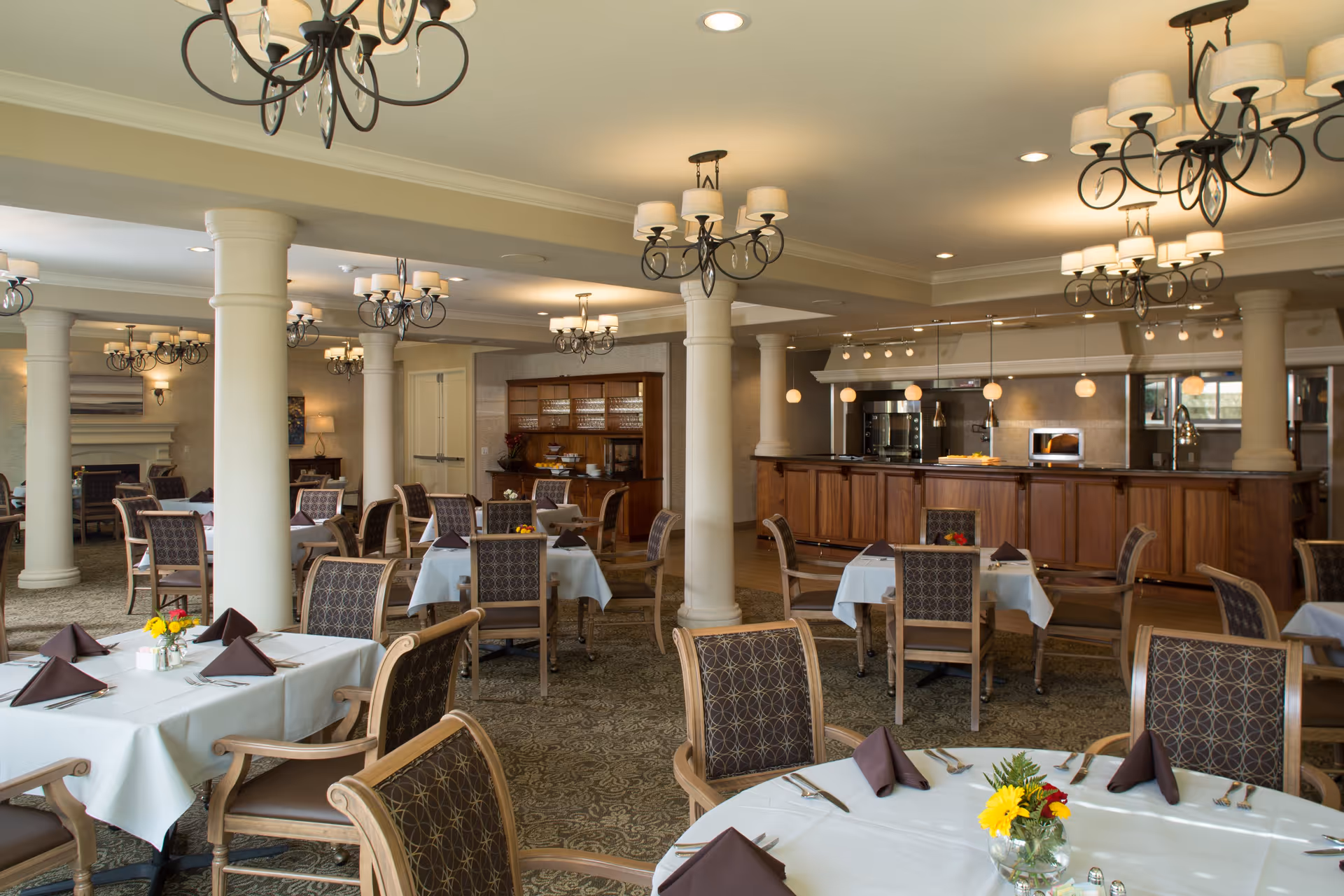A spacious dining room with multiple round and square tables covered with white tablecloths, each set with brown folded napkins, silverware, and small floral centerpieces. The room features ornate chandeliers, large white columns, patterned carpet, and a wooden buffet area with kitchen appliances in the background.