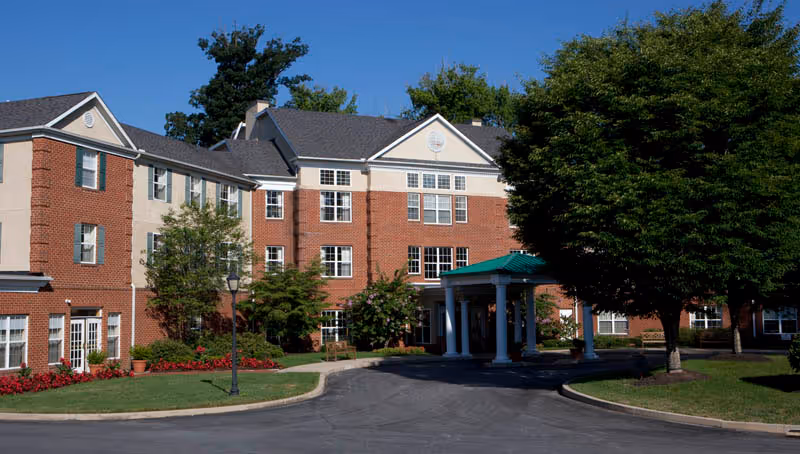 Brick three-story senior living building with a covered entrance, driveway, trees, and landscaped beds under a clear blue sky.