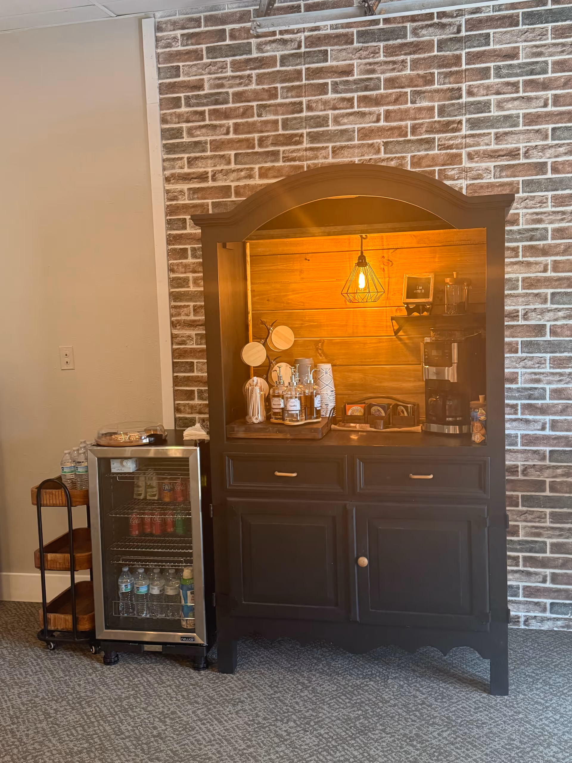 Dark wood coffee bar with warm lighting against brick wall beside a small beverage fridge.