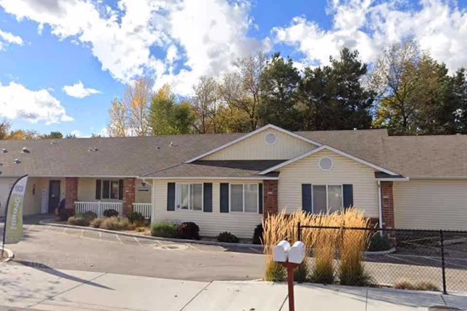 A single-story beige senior care facility with brick accents, a driveway, and a fenced front yard.