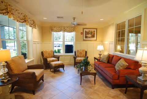 Sunlit communal living room with a red sofa, armchairs, a coffee table, and large windows.