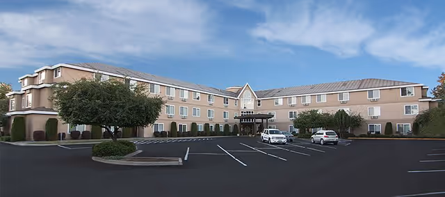 Exterior view of a three-story senior living facility building with a large parking lot in front, a few parked cars, and some trees and shrubs around the building under a partly cloudy sky.