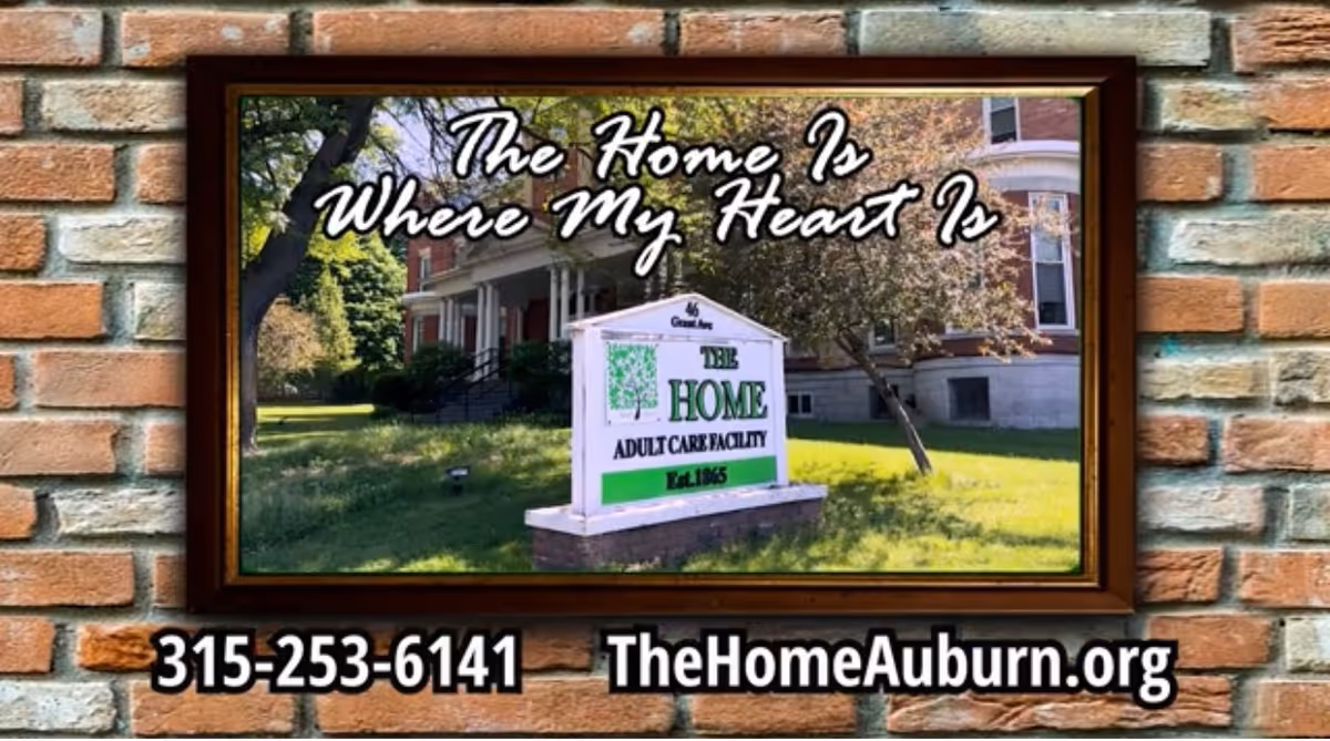 A framed photo on a brick wall showing the exterior of an adult care facility with a sign that reads 'THE HOME ADULT CARE FACILITY Est. 1865'. Above the photo, text says 'The Home Is Where My Heart Is'. Below the frame, there is a phone number 315-253-6141 and a website TheHomeAuburn.org.