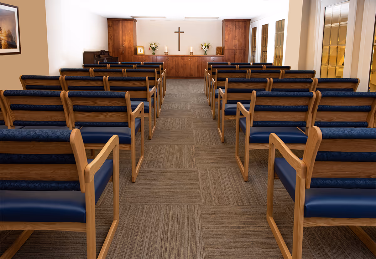 Interior view of a chapel with rows of wooden pews with blue cushions facing a wooden altar. The altar has a cross mounted on the wall above it, two lit candles, flower arrangements, and a framed picture. There is a piano on the left side and stained glass windows on the right side.