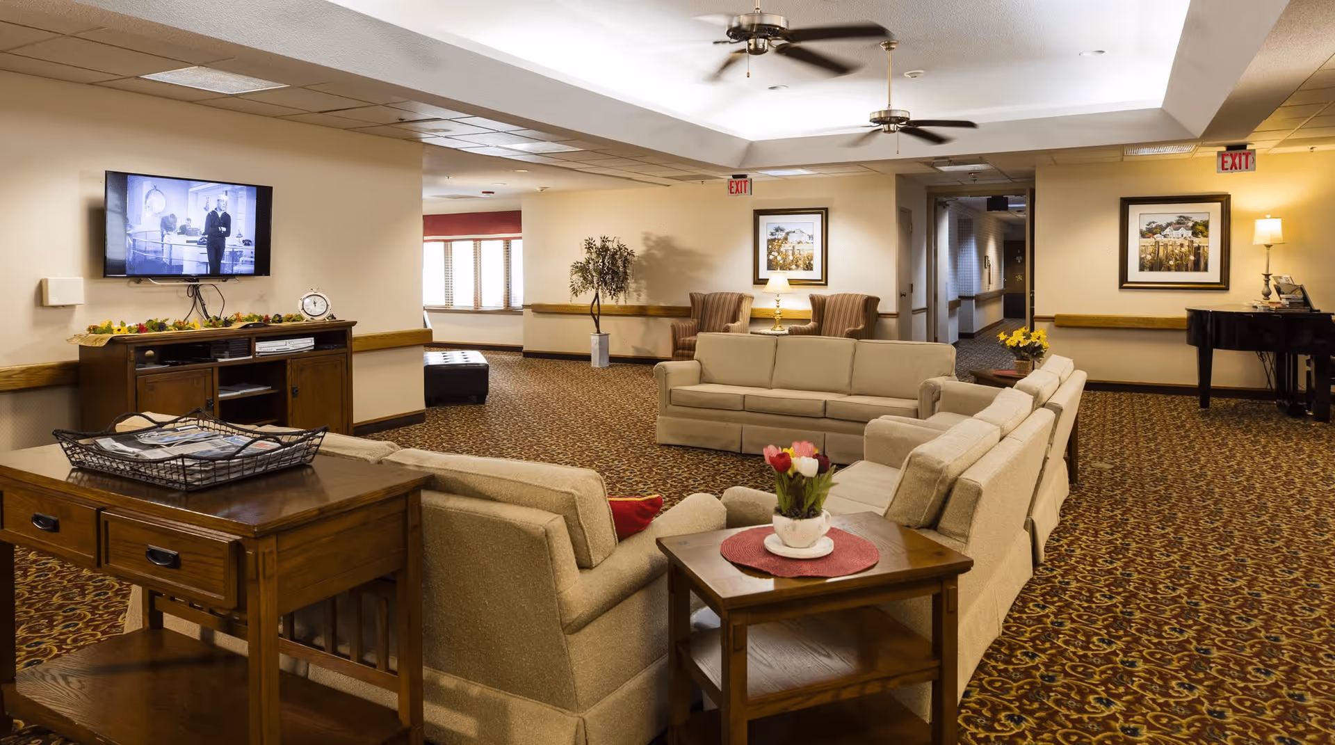 A spacious living room area in a senior living facility with beige sofas arranged around wooden side tables, a TV mounted on the wall showing a black and white program, two armchairs with a lamp and framed artwork on the wall, a piano in the corner, ceiling fans, and patterned carpet flooring.