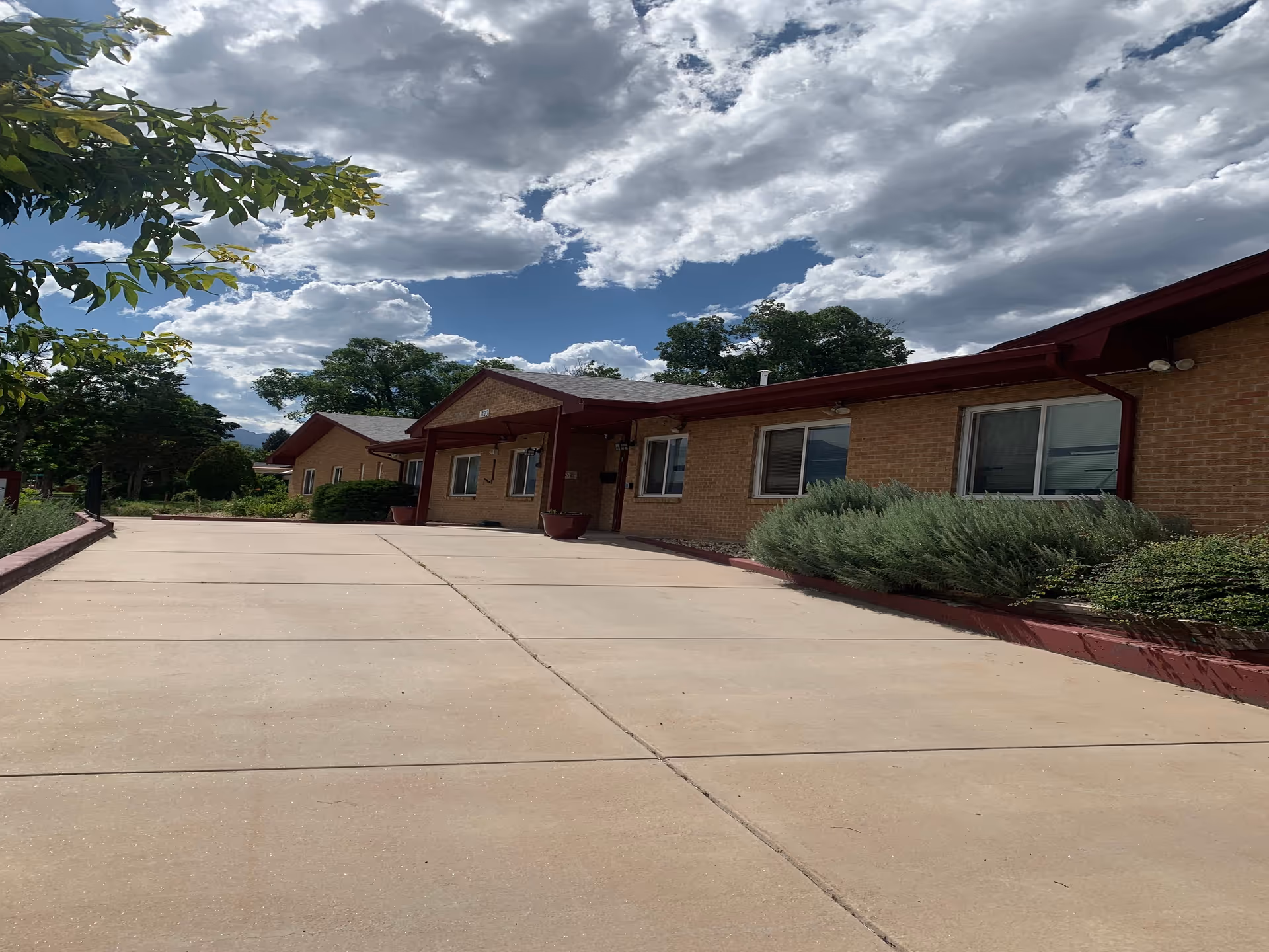 Single-story brick building with a concrete driveway, shrubs, and cloudy sky above.