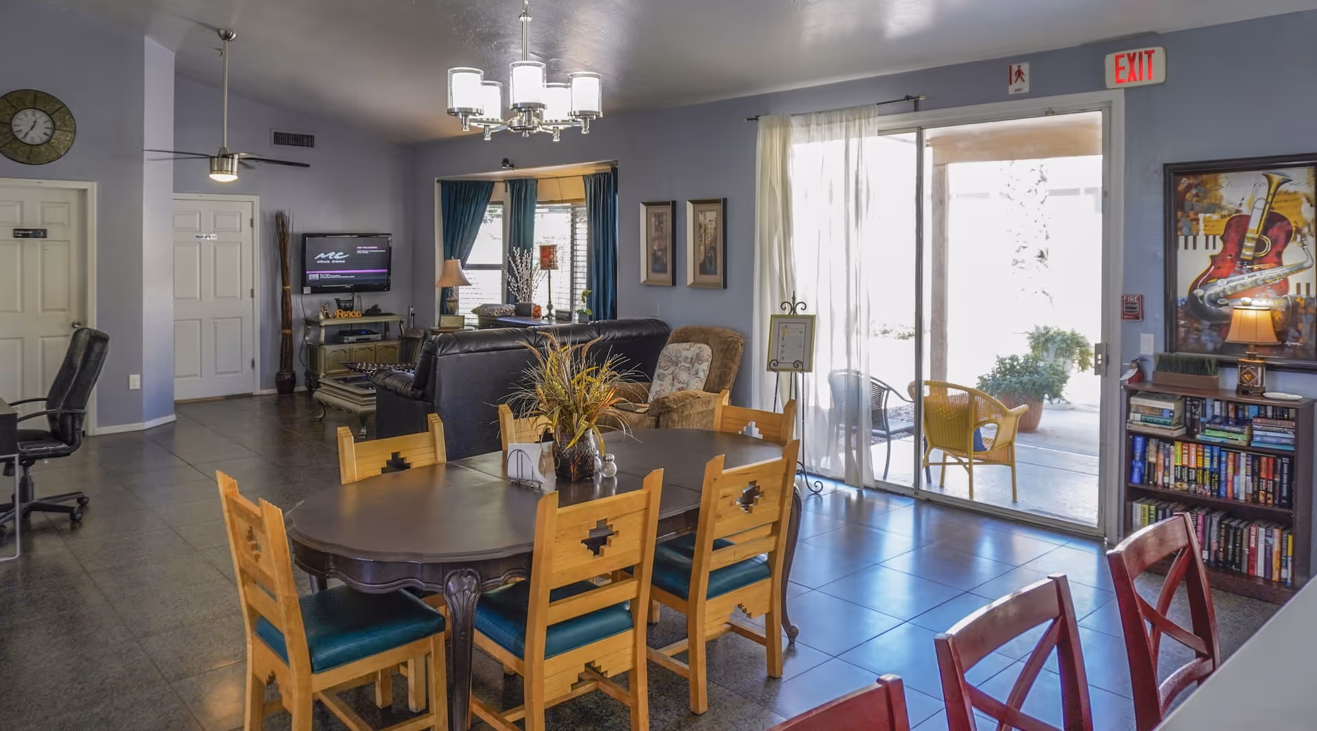 A bright and spacious common area in an assisted living home featuring a wooden dining table with six chairs, a black leather sofa, an armchair, a TV mounted on the wall, and a bookshelf filled with books. Large sliding glass doors lead to an outdoor patio with chairs and plants. The room has light blue walls, tile flooring, and decorative artwork on the walls.