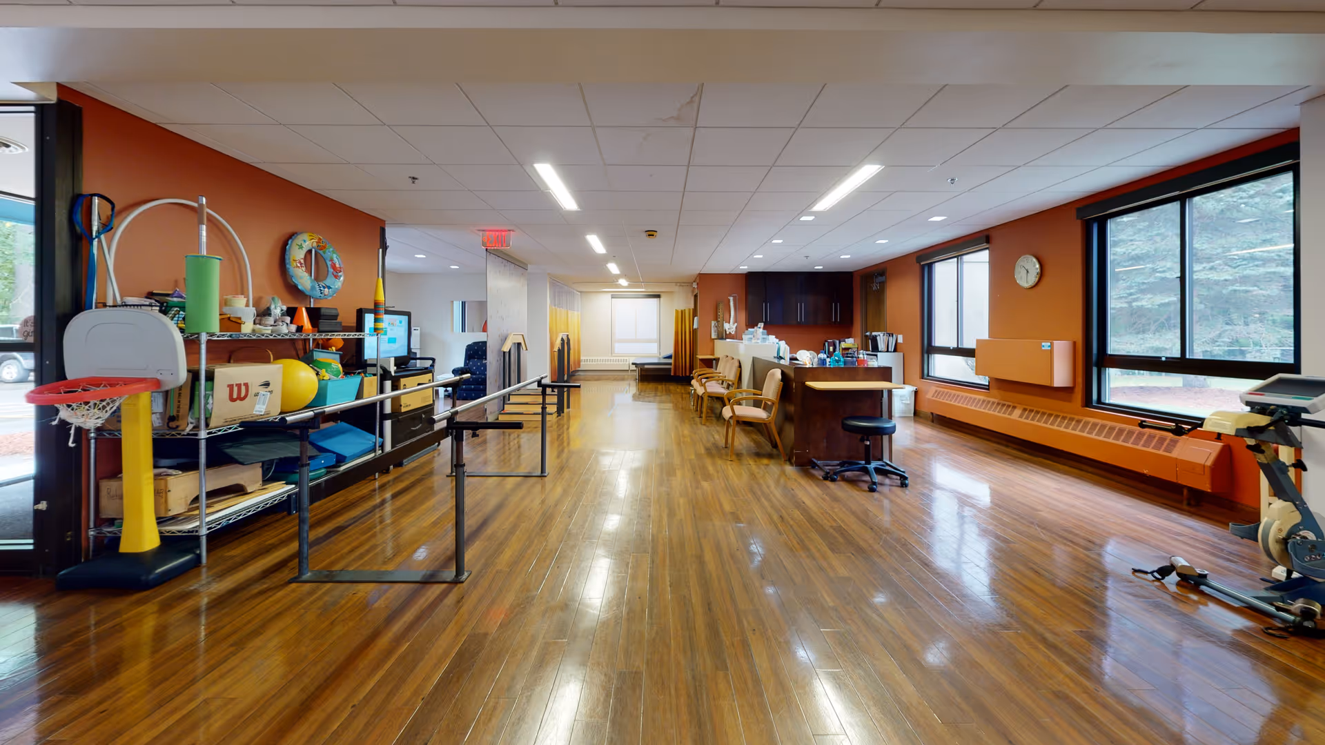 A spacious room with polished wooden floors and large windows letting in natural light. The room contains exercise and therapy equipment including parallel bars, a small basketball hoop, a stationary bike, and various therapy supplies on shelves. There are chairs and a counter area along one wall, with orange walls and a clock mounted near the windows.
