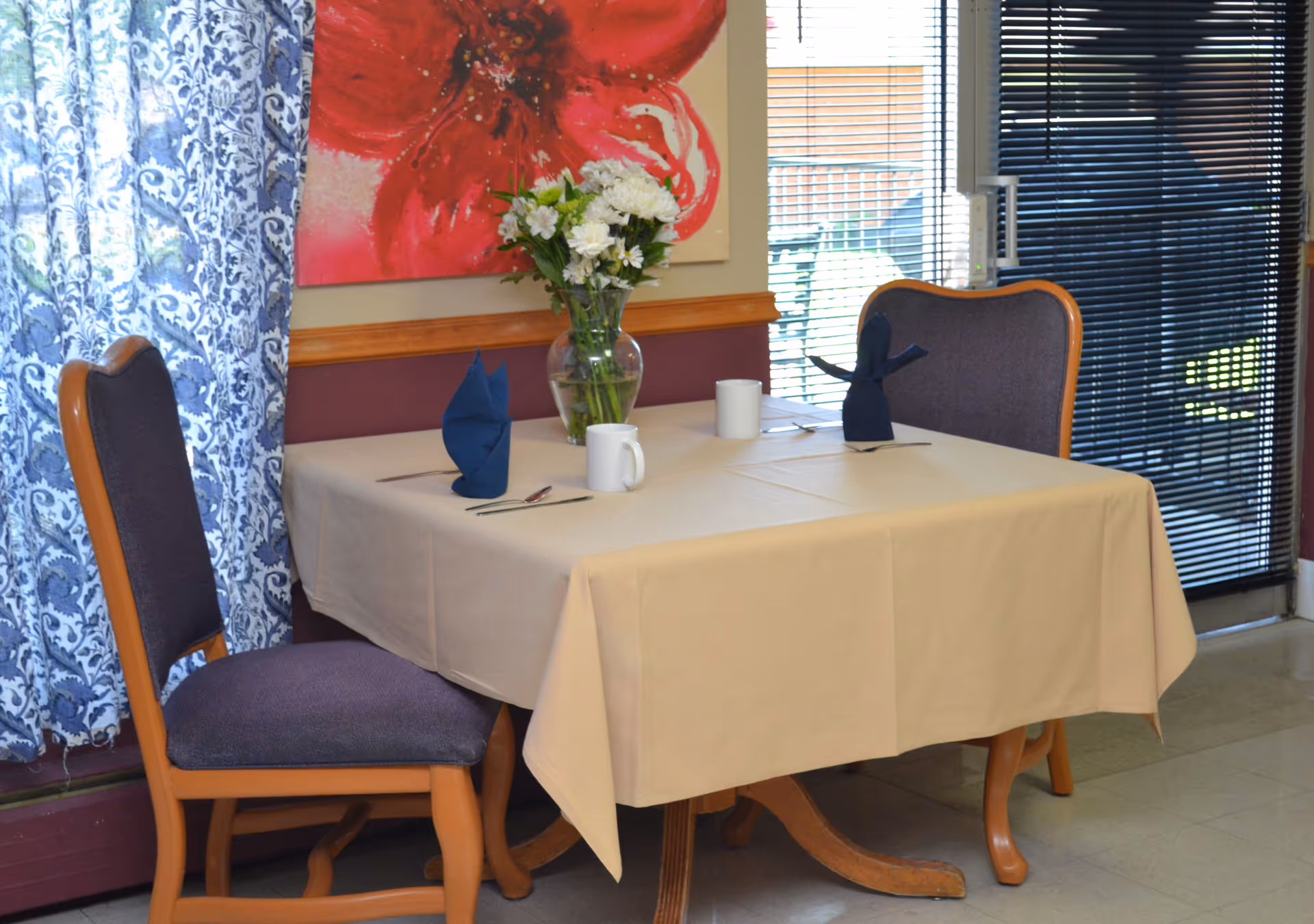 A small dining table set for two with a beige tablecloth, two purple cushioned chairs, two white mugs, silverware, and blue folded napkins. A vase with white flowers is placed in the center of the table. Behind the table is a large red flower painting and a window with blue patterned curtains on the left and black blinds on the right.