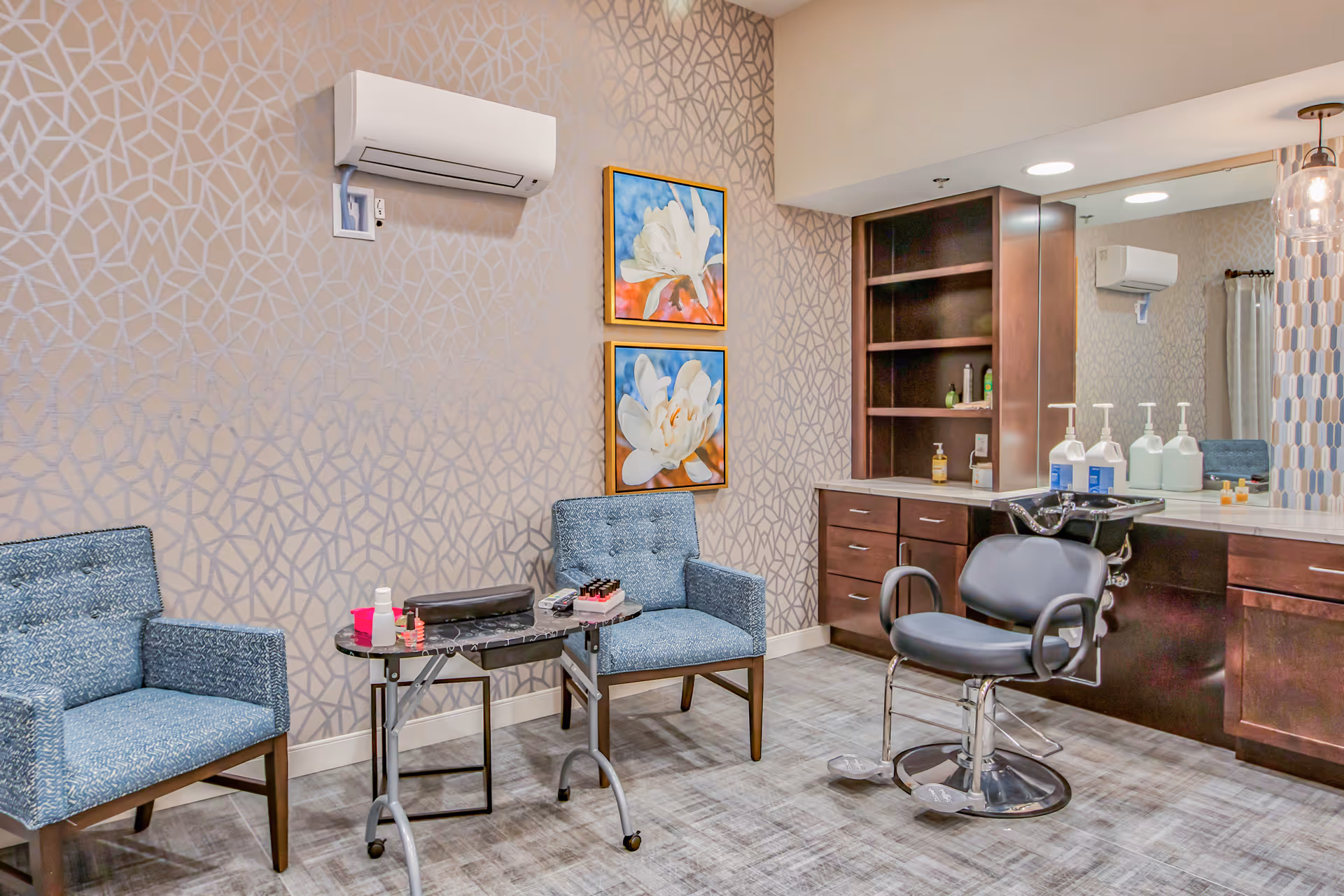 A salon area in a senior living facility with two blue upholstered chairs, a small table with nail polish and manicure supplies, a black salon chair in front of a counter with multiple bottles of lotion or sanitizer, wooden cabinets, a large mirror, and two floral paintings on the wall under a mounted air conditioning unit.