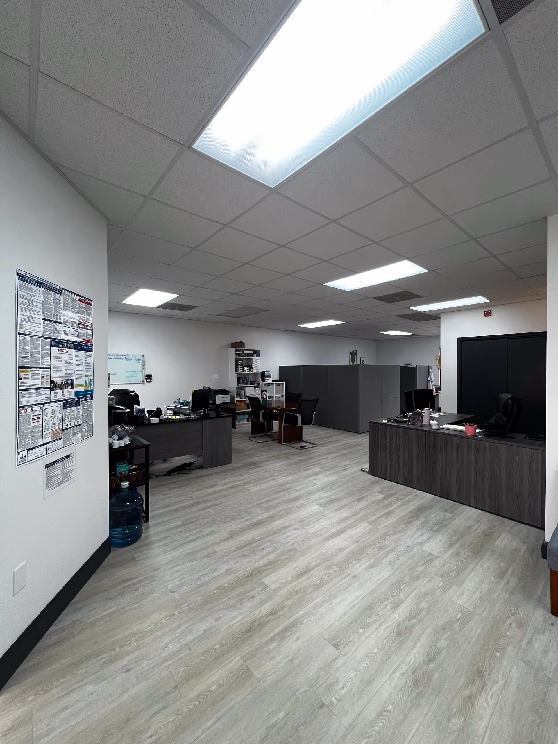 Bright office area with desks, chairs, cubicles, and informational posters.