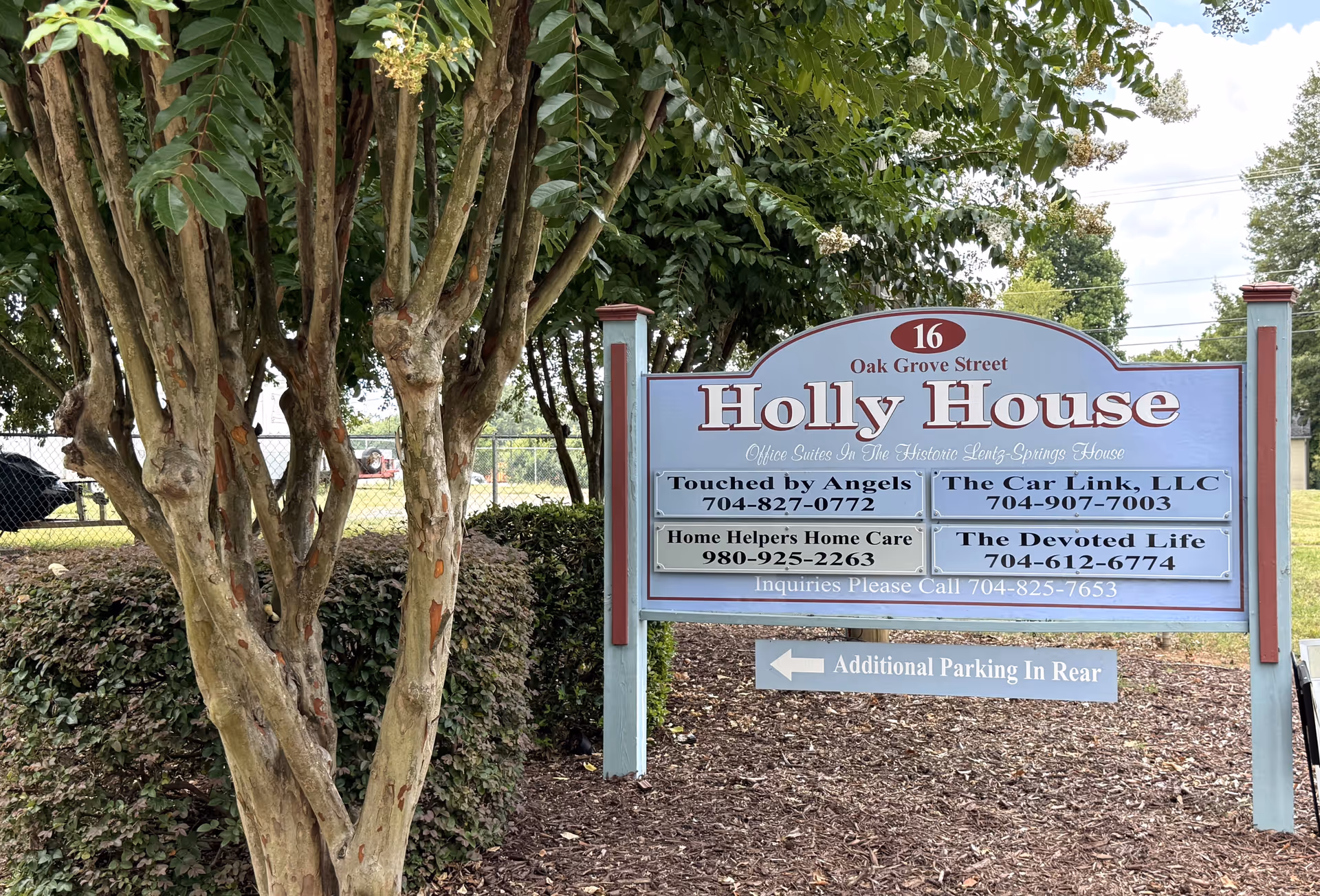 Outdoor signboard for Holly House with business names, trees, and a parking area.
