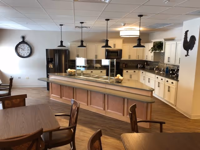 A spacious kitchen and dining area with wooden tables and chairs in the foreground. The kitchen features white cabinets, a black refrigerator, a microwave, and a large island with a protective glass barrier and decorative bowls. A large wall clock and a rooster decoration are visible on the walls.