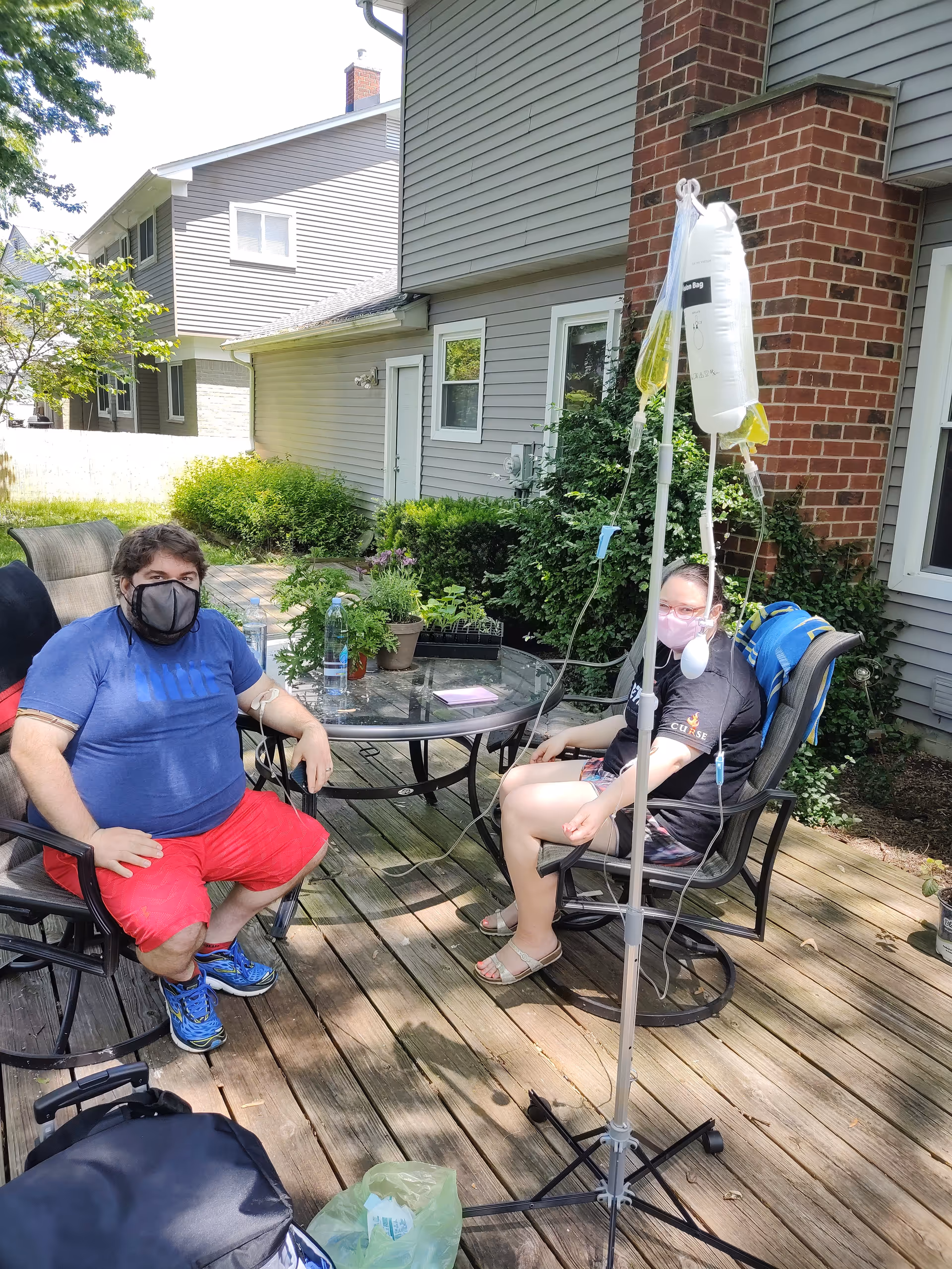 Two residents wear masks while sitting outside on a wooden deck with an IV stand nearby.