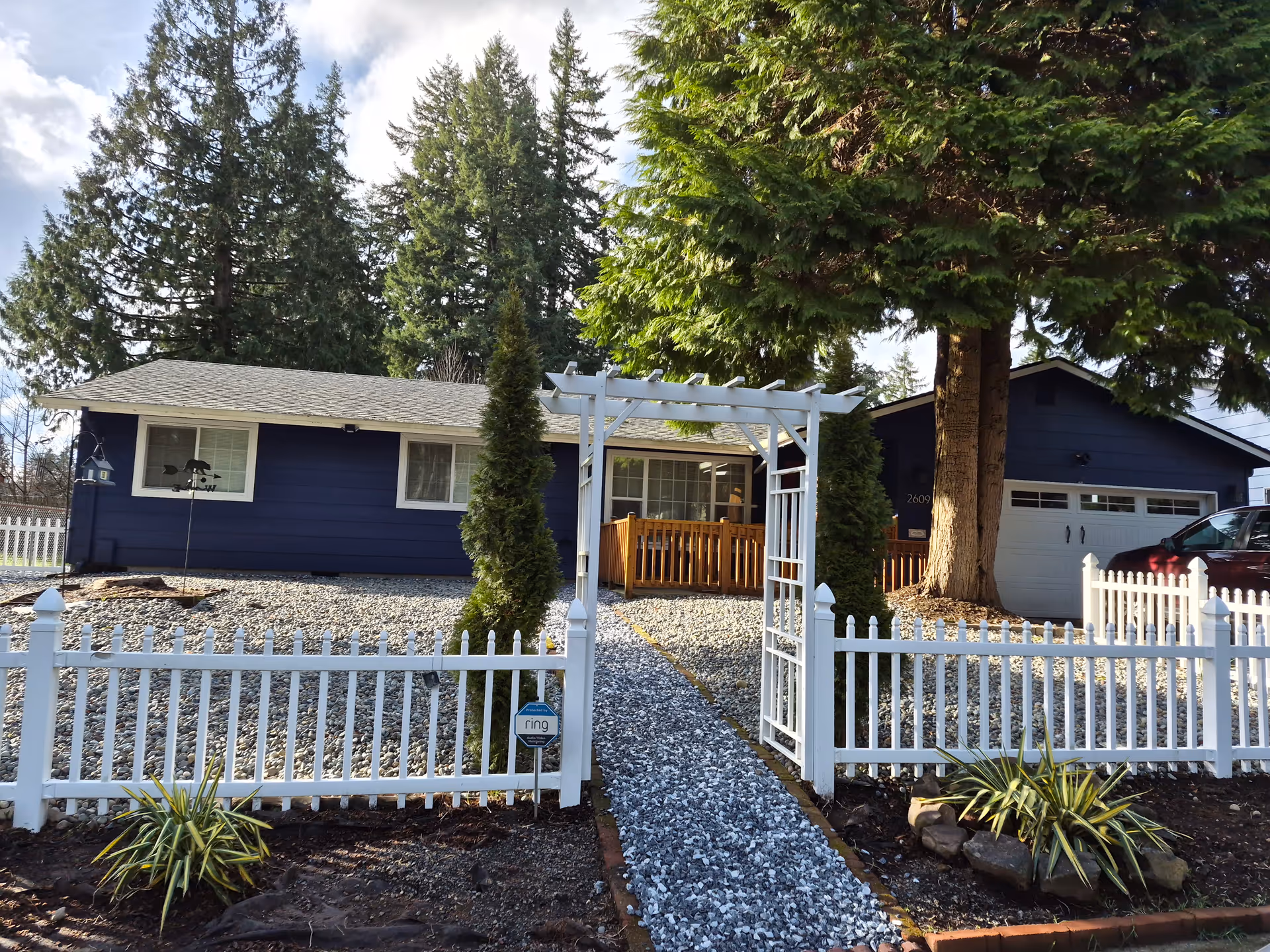 Blue single-story senior care home with white picket fence, arch gate, and gravel pathway.