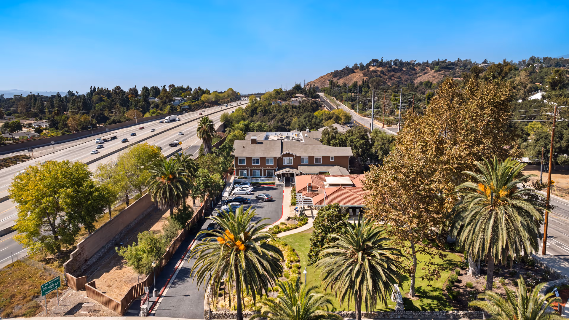 Aerial view of Ivy Park at Claremont facility showing a large building surrounded by palm trees and other greenery. There is a parking lot with several cars, a well-maintained lawn, and a highway with vehicles visible to the left. Hills and a clear blue sky are in the background.