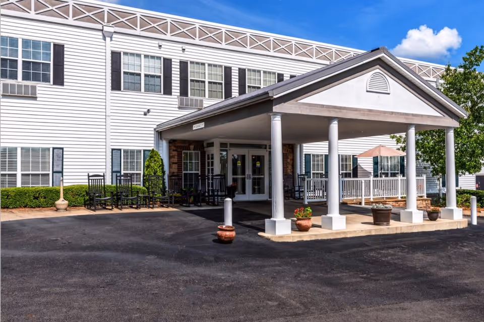 Covered entrance and porte-cochère of a white multi-story senior living building with rocking chairs, potted plants, and a driveway.