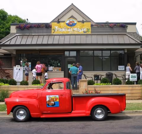 Exterior view of a building with a sign reading 'Tropical Moose' above the entrance. Several people are standing outside near the entrance and patio area with tables and chairs. A bright red vintage pickup truck with a moose logo on the door is parked in front of the building.