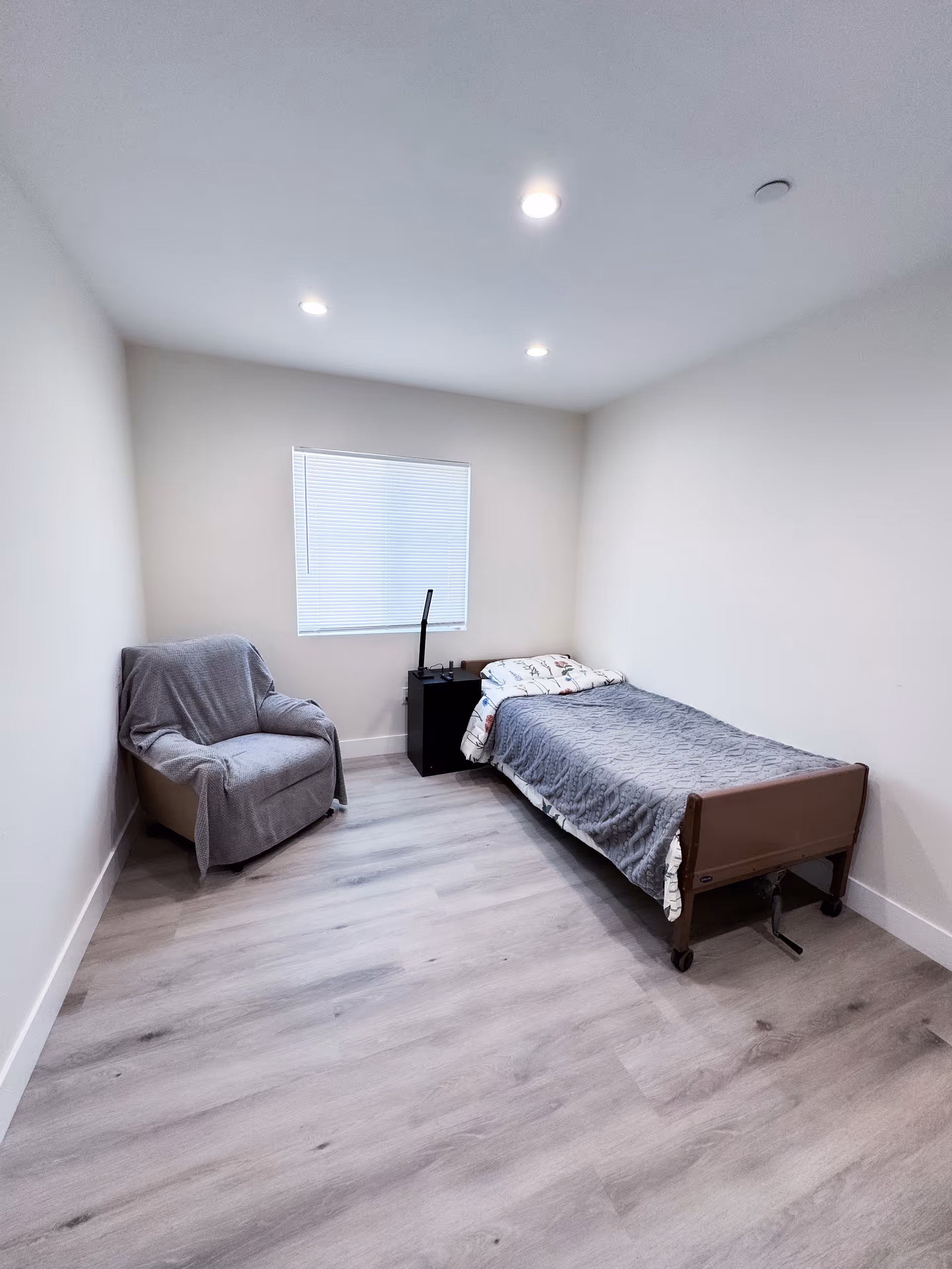 A small, modern bedroom with a single bed, armchair, nightstand, and window blinds.