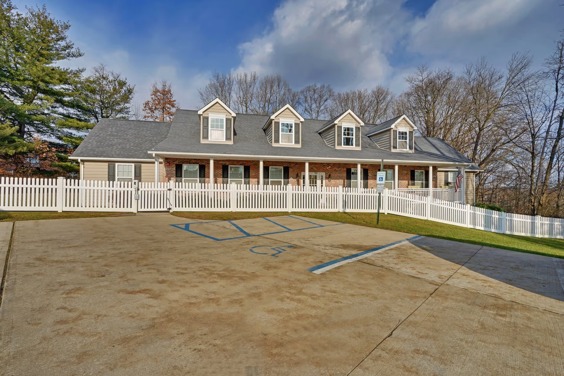 Exterior view of a single-story brick and siding building with a gray roof and four dormer windows. The building is surrounded by a white picket fence and has a concrete parking area in front with marked handicap parking spaces. Trees without leaves are visible in the background under a partly cloudy sky.