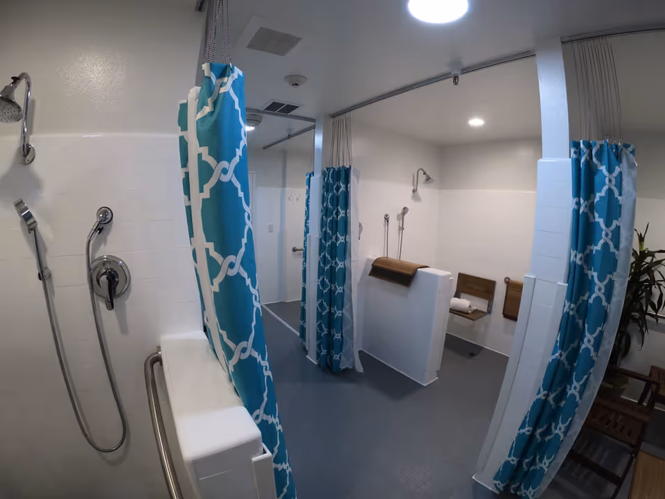 Accessible communal shower area with teal patterned curtains, handheld showerheads, fold-down seats and grab bars.