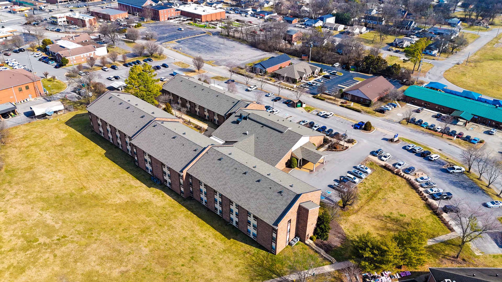 Aerial view of Park Place Retirement Community showing a large brick building with a gray roof surrounded by parking lots, grassy areas, and nearby streets with other buildings and trees in the background.