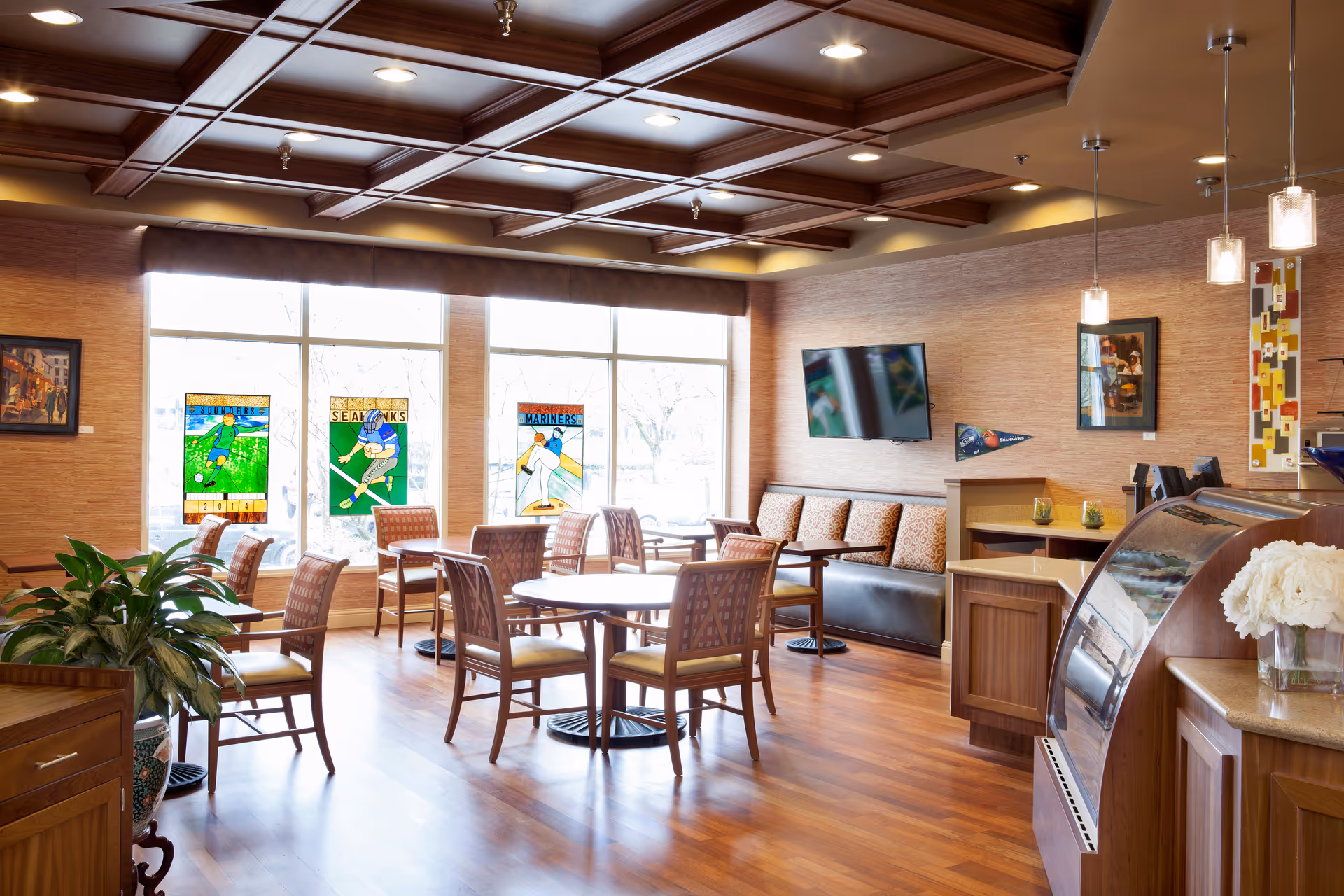 A cozy senior living lounge with tables, chairs, large windows, and a curved service counter.