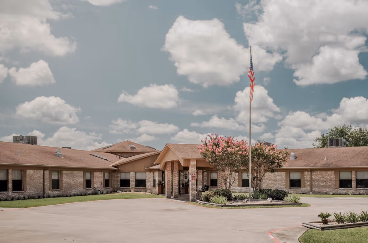 Exterior view of Southbrooke Manor Nursing and Rehabilitation Center showing a single-story brick building with a peaked entrance, an American flag on a flagpole, and flowering trees in front under a partly cloudy sky.