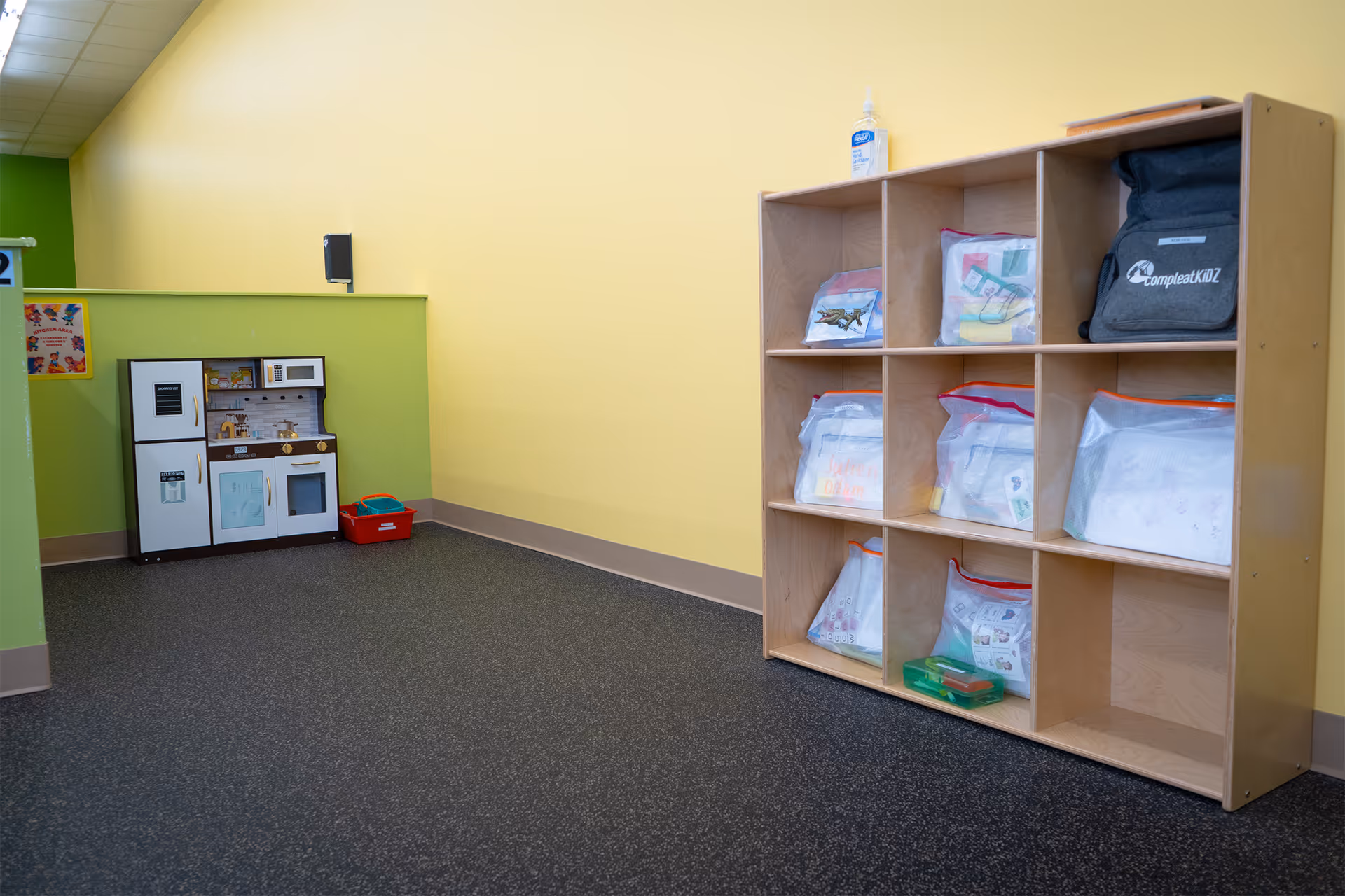 Bright playroom with a wooden cubby shelf filled with labeled bags and a toy kitchen.